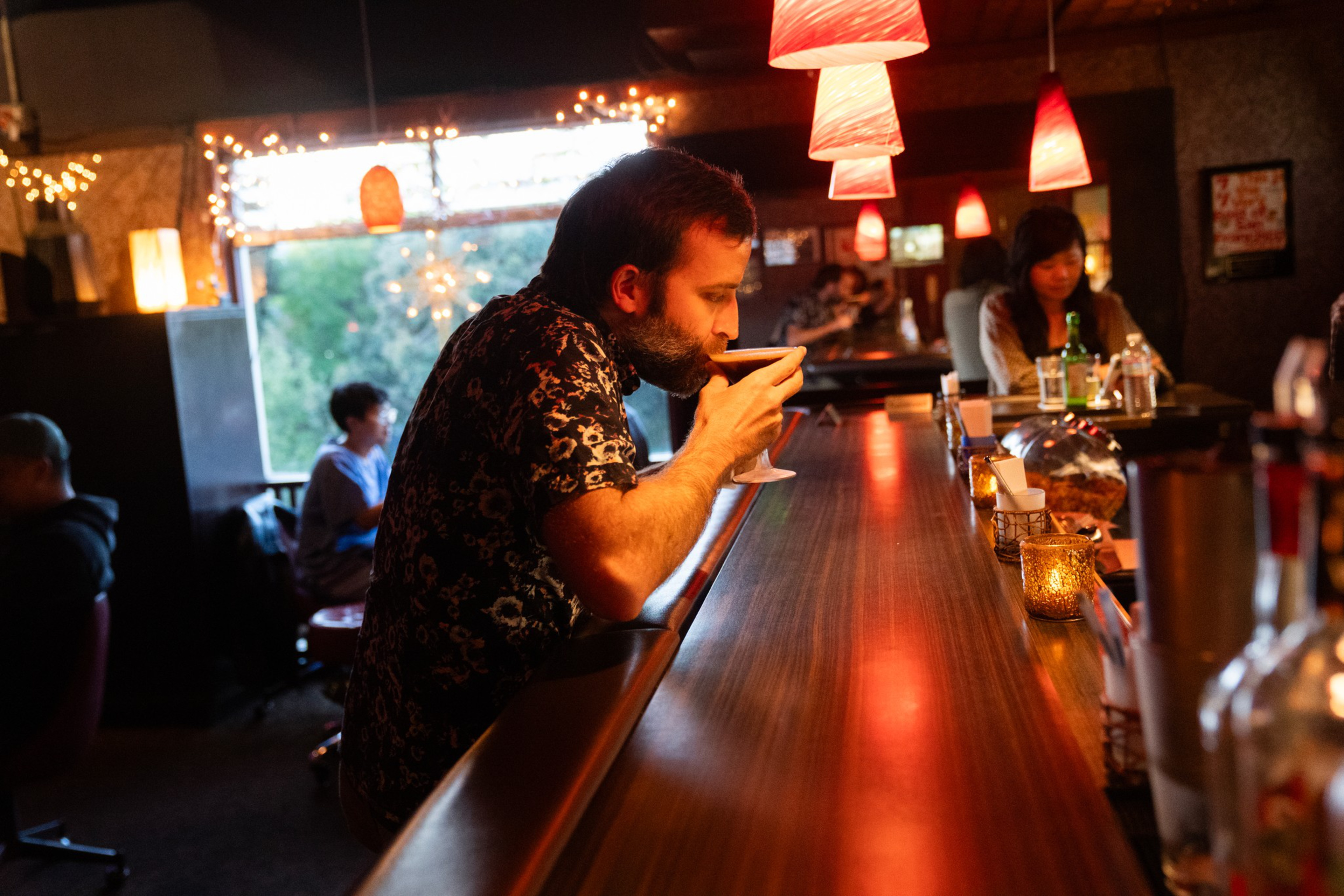 A man with a beard drinks from a cocktail glass at a wooden bar counter lit by red hanging lamps, with other patrons in a dimly lit room.