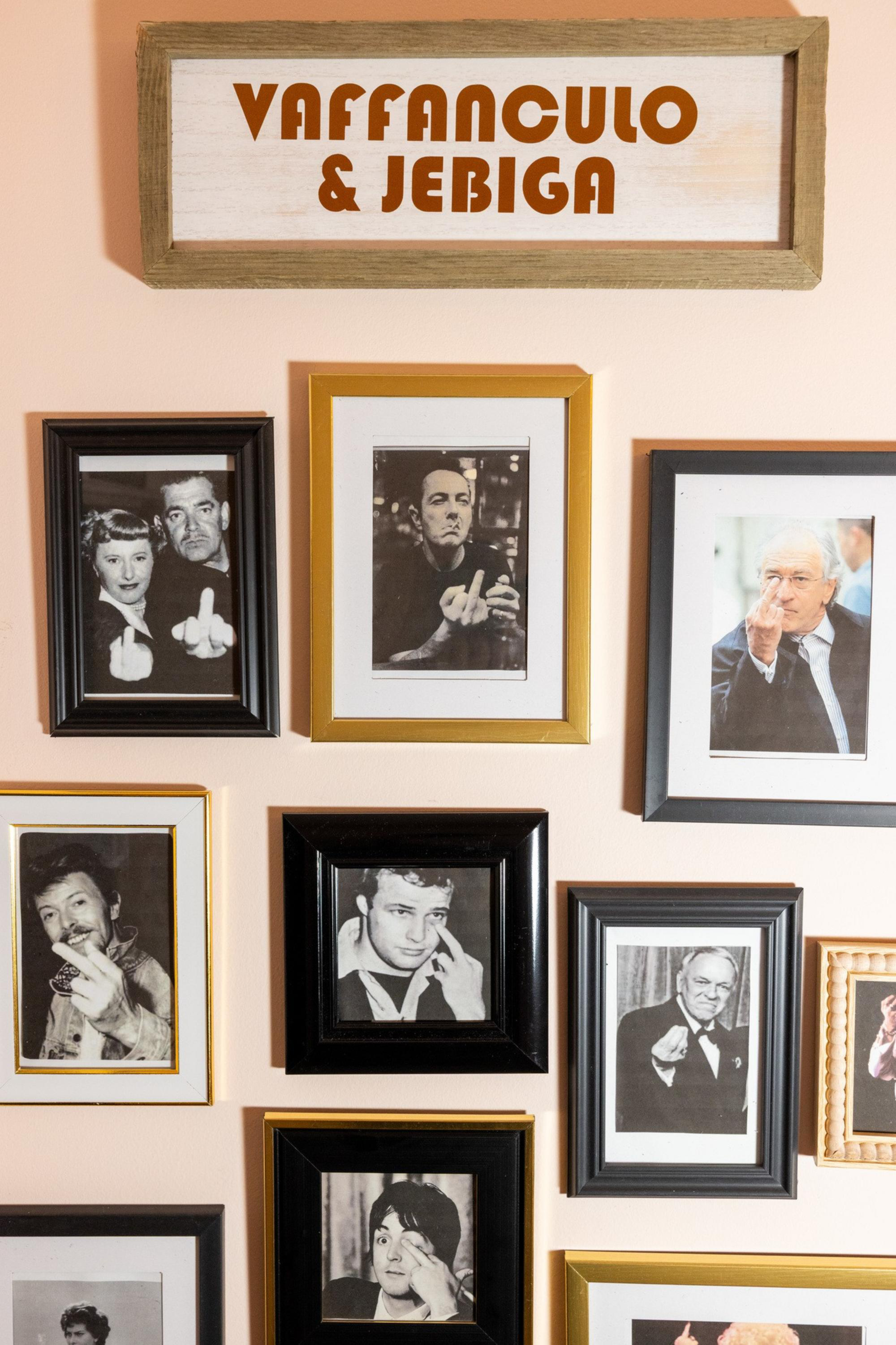 A collection of framed black-and-white and color photos features various people giving the middle finger, with a sign above reading “Vaffanculo & Jebiga.”