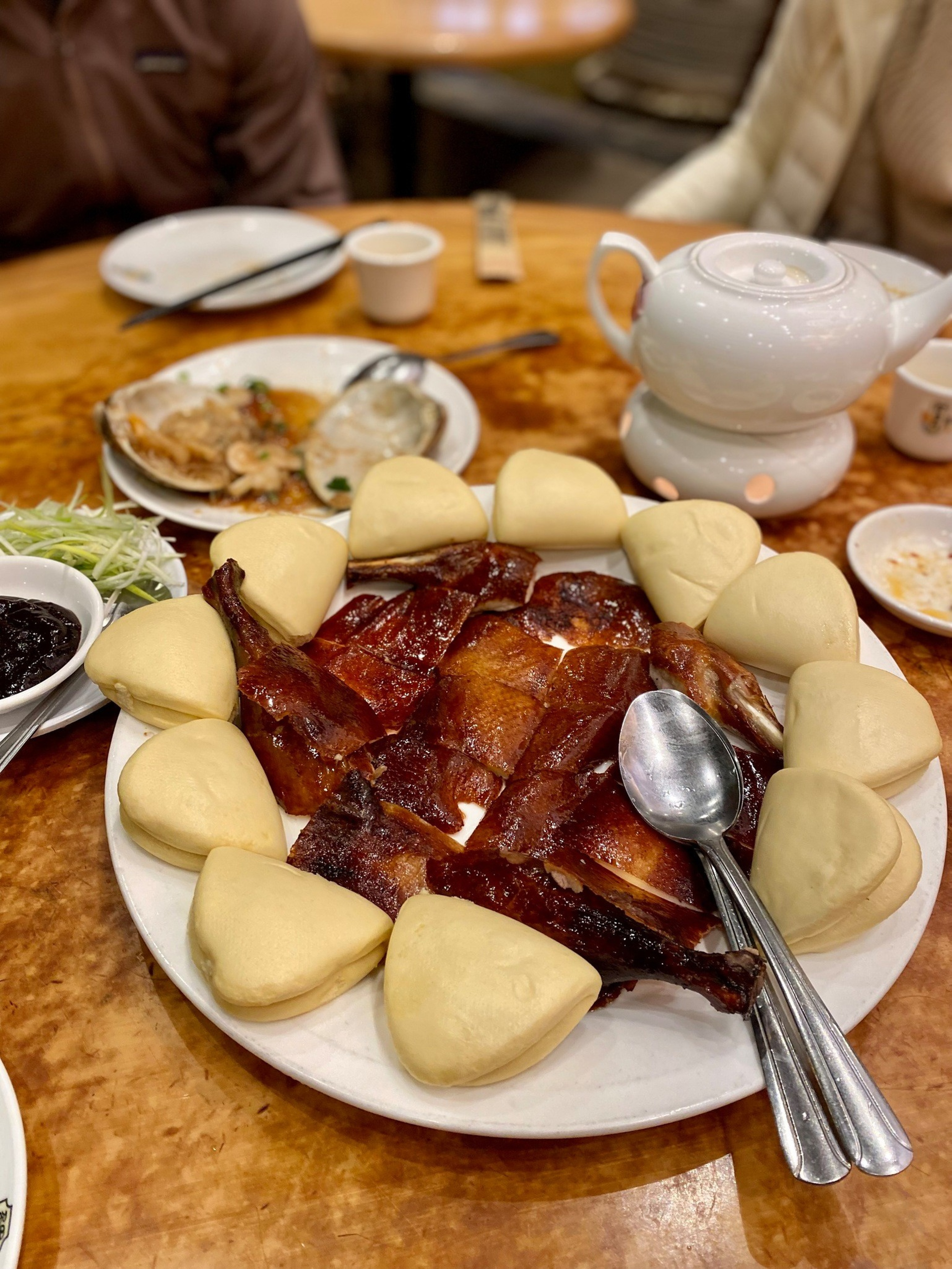 Roast duck pieces are arranged on a plate surrounded by steamed buns, with two spoons resting on the plate, next to a teapot and other dishes.