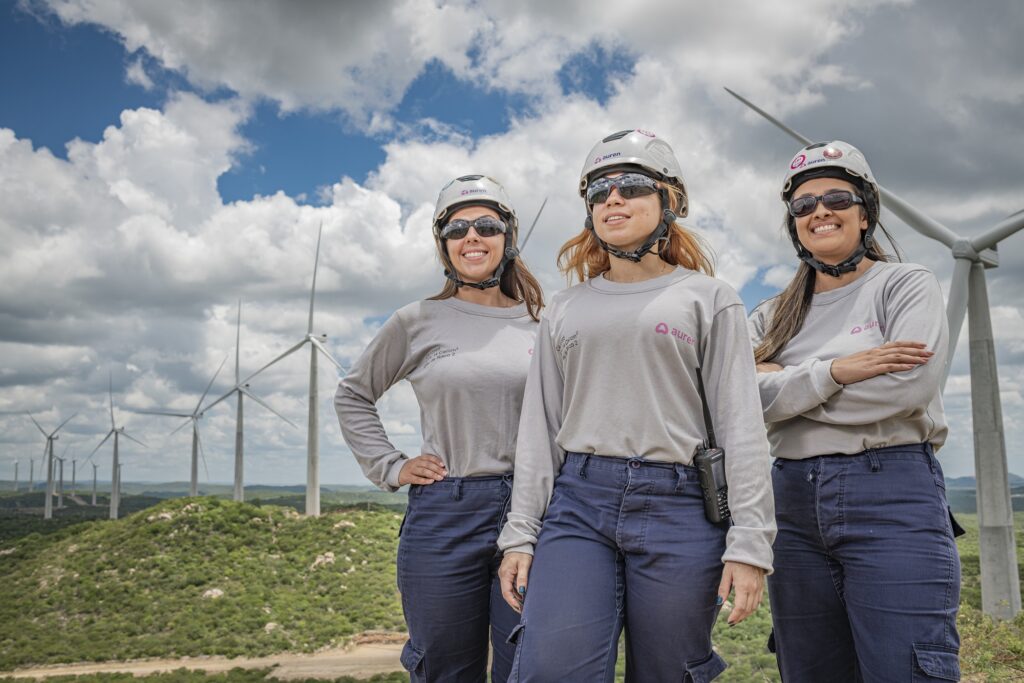 Three women, all wearing hard hats and matching gray shirts, stand in front of a group of wind turbines.