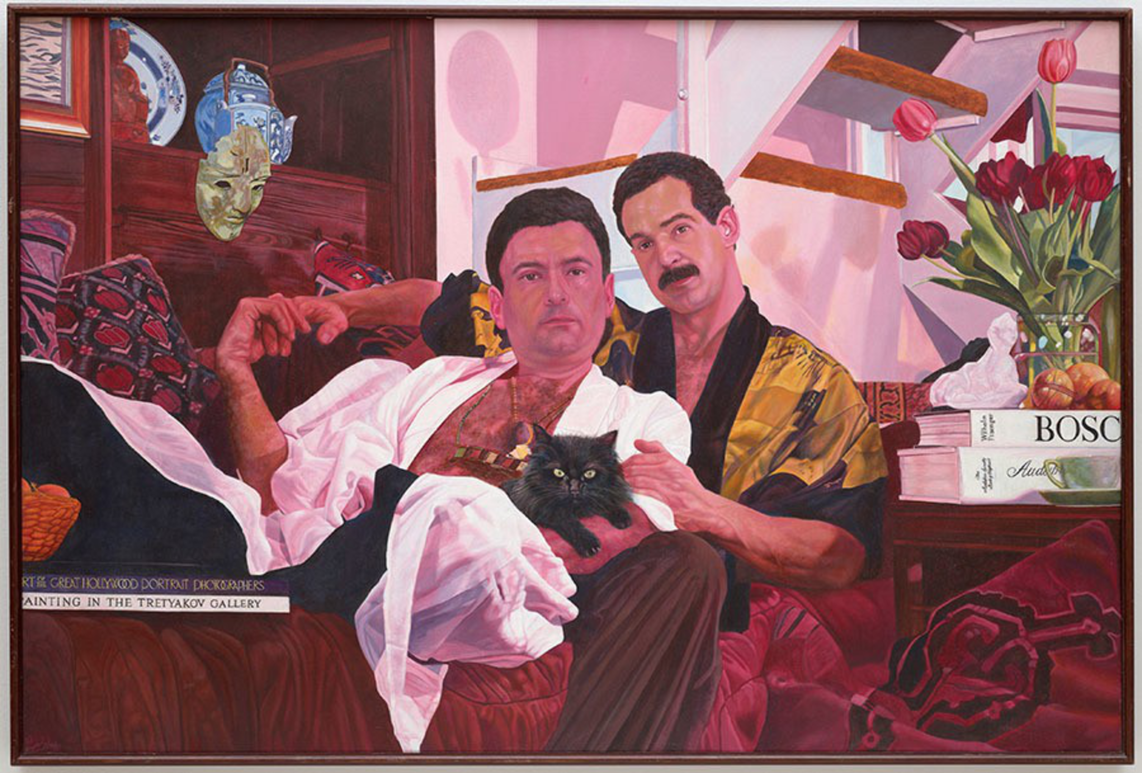 Two men sit closely on a couch, one holding a black cat, with a wooden mask, tulips, and books on a side table nearby.