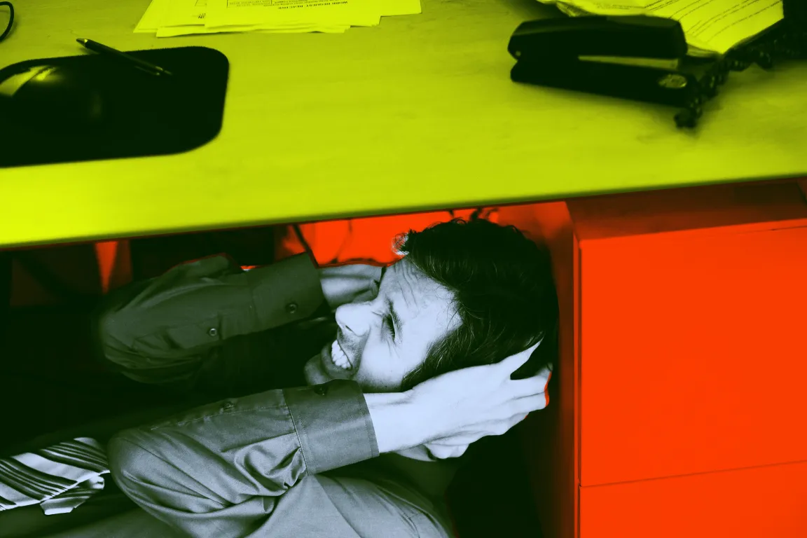 A man is crouched under a desk, covering his ears with his hands and grimacing, suggesting distress or discomfort. The desk surface above him is cluttered with papers, a stapler, a telephone, and a pen on a mouse pad. The image has a color filter with yellow on the desk, red on the background and drawer, and a greenish tint on the man and his clothing.