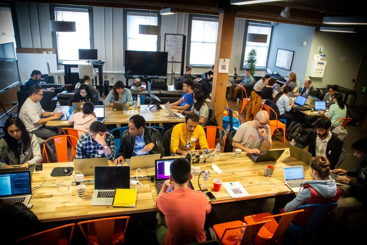People sitting around large tables, working on their laptops, and talking in a crowded room.