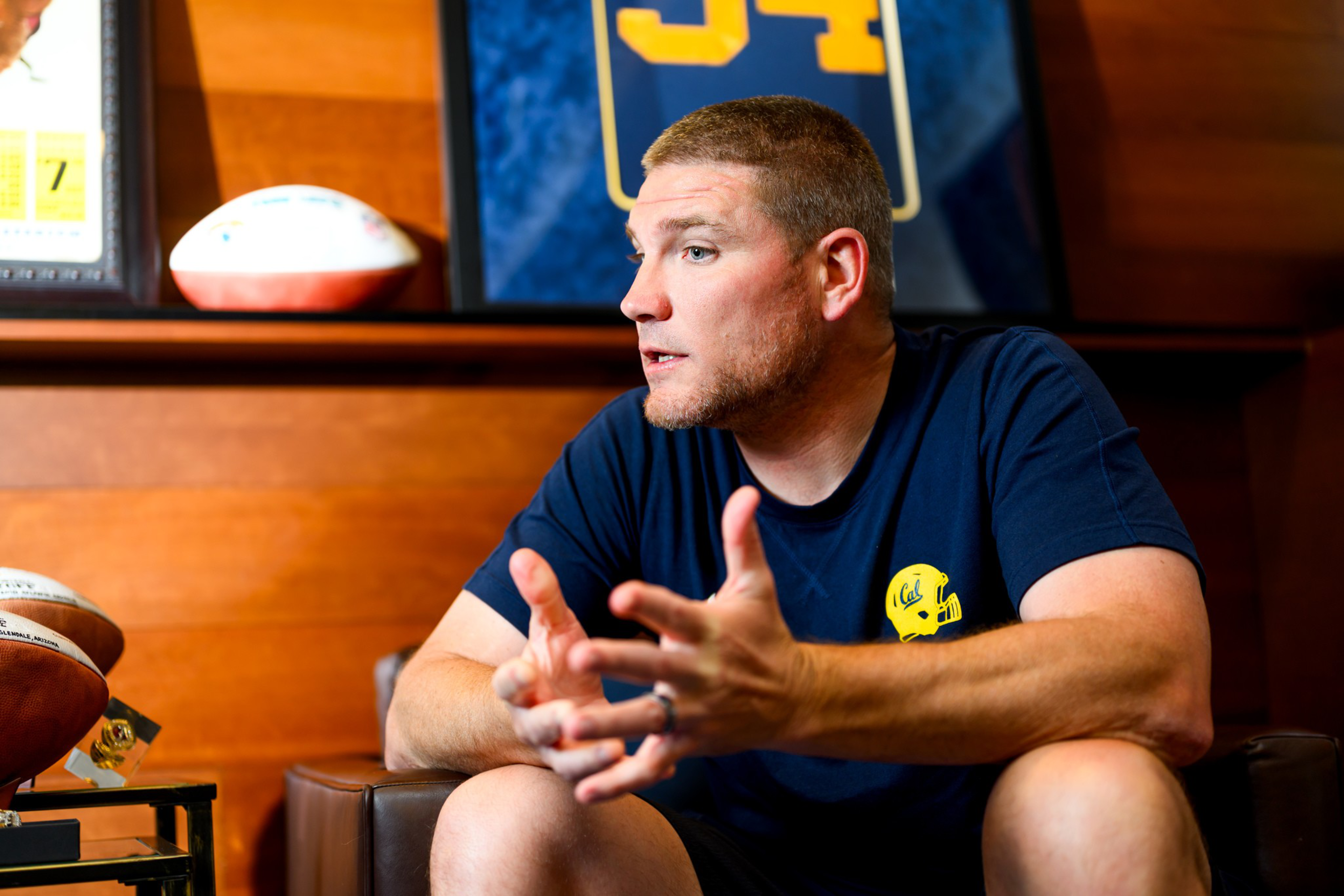 A man in a navy blue shirt with a yellow helmet logo talks animatedly while sitting, with framed sports memorabilia and footballs behind him.
