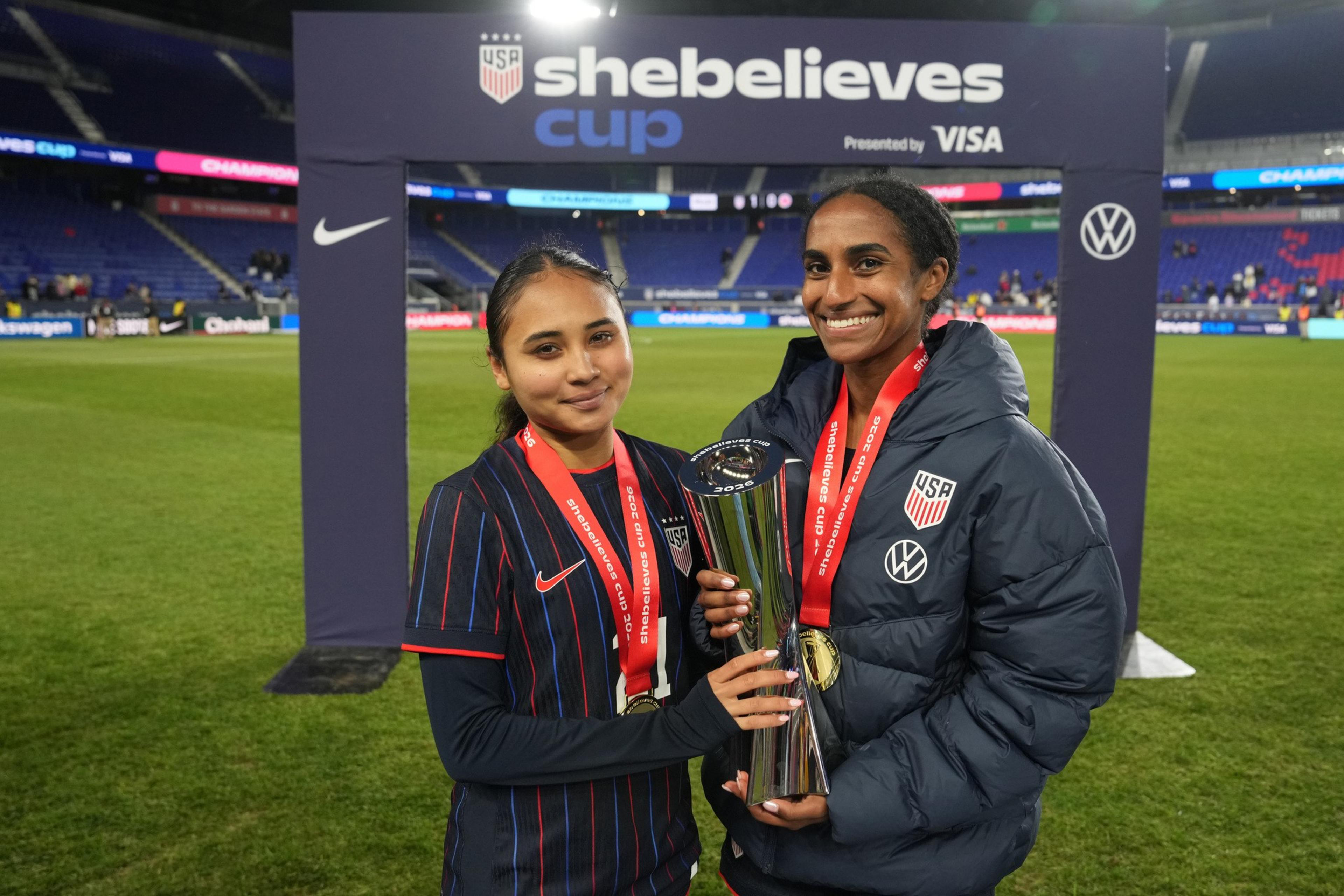 Two female soccer players wearing USA team gear smile while holding a silver trophy with red medal ribbons around their necks on a soccer field.