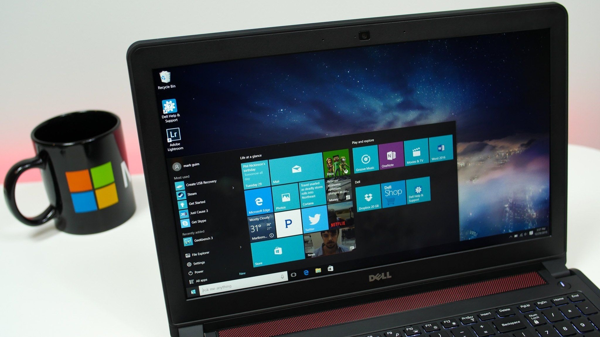 A Dell laptop showing the Windows start menu with a galaxy-themed wallpaper. To the left, a black mug with colorful square logos sits on a white table.