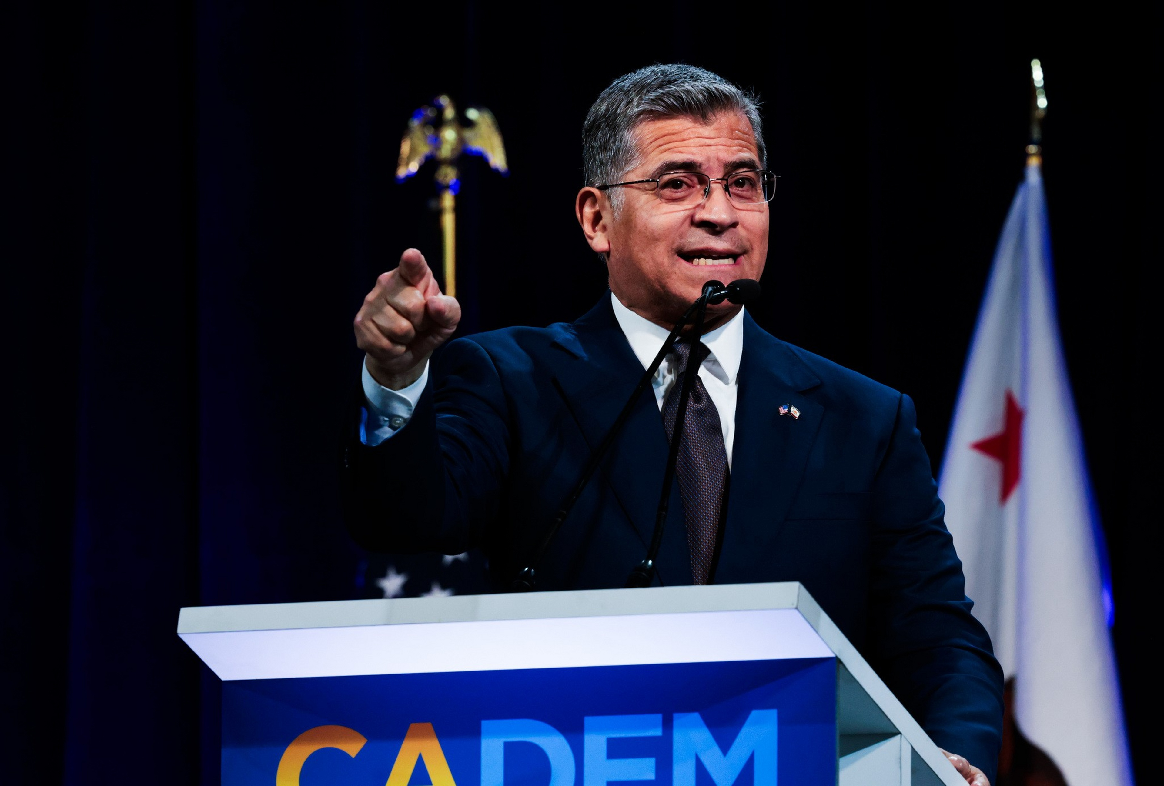 A man in a dark suit and glasses speaks at a podium labeled “CADEM,” pointing with his right hand, with an American flag and another flag behind him.
