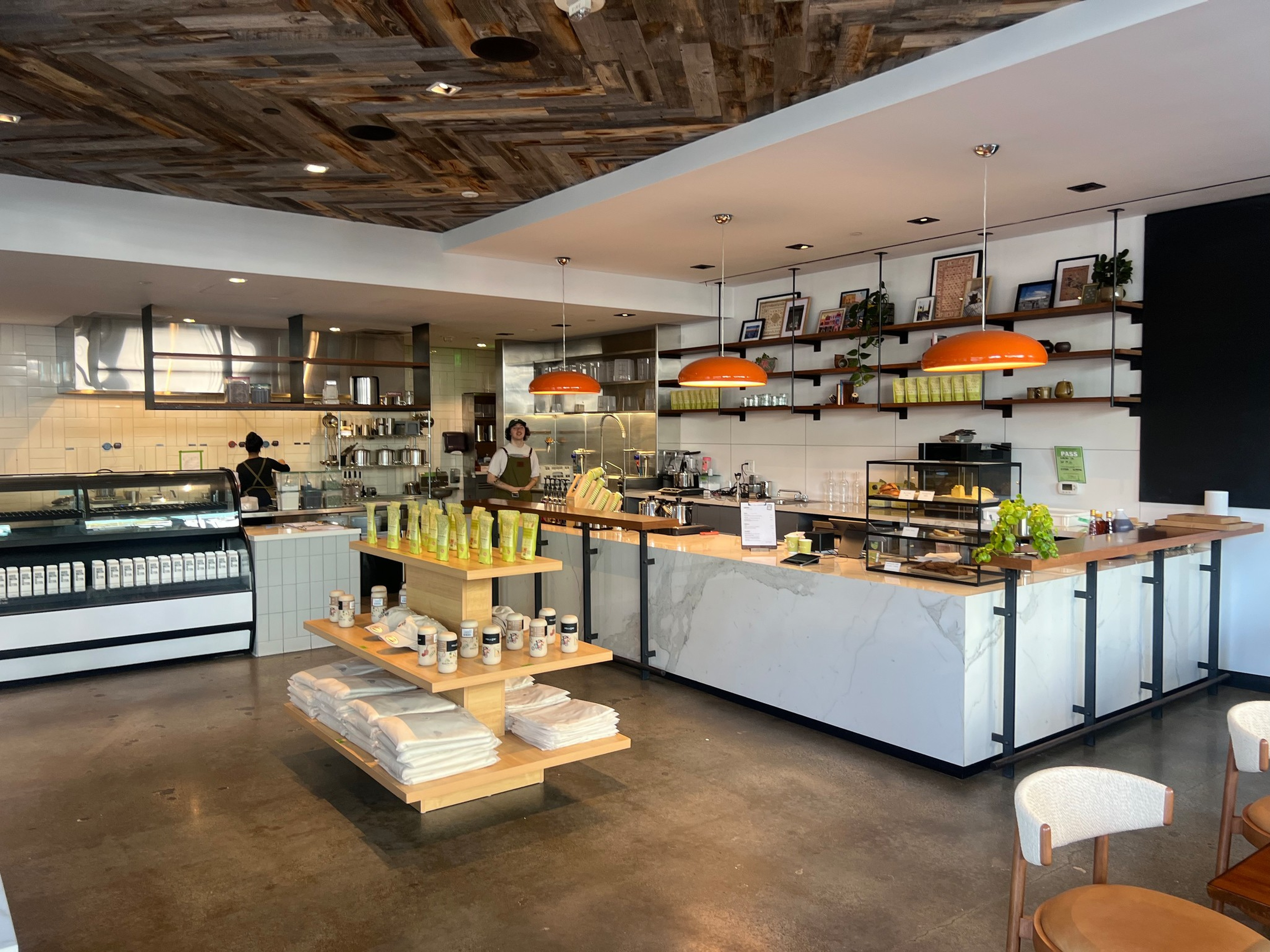 A modern café interior with a wooden ceiling, orange pendant lights, a marble counter, shelves with products, and two staff members behind the counter.