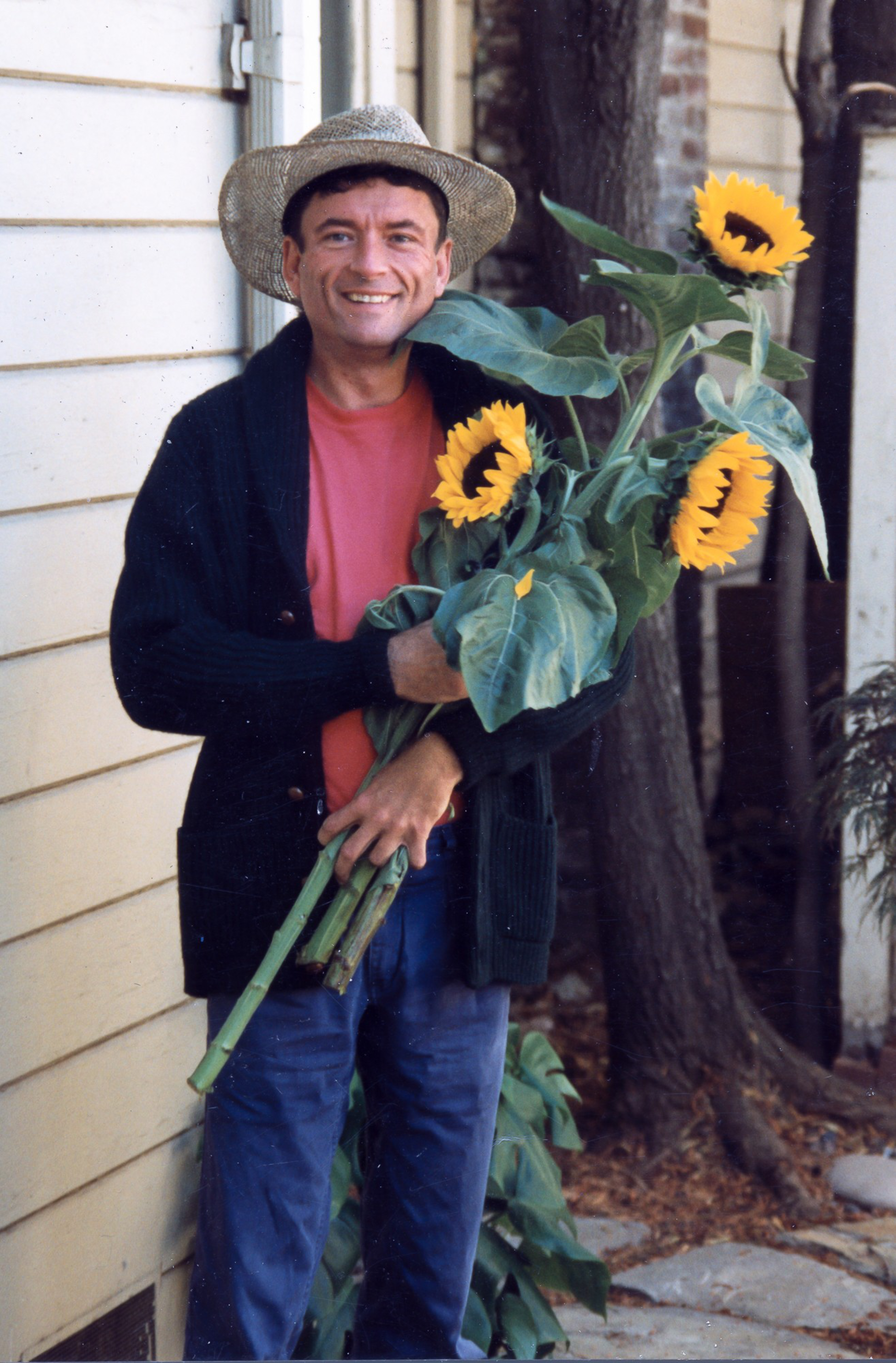A man in a straw hat, red shirt, dark cardigan, and blue pants smiles while holding three large sunflowers outside near a cream-colored wall and tree.