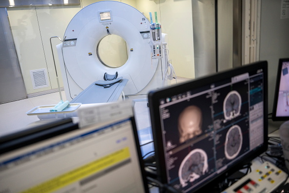 A look inside the analysis room next to a large white circular machine, or the CT machine. Inside the room a computer monitor shows four side by side images of a brain being scanned.