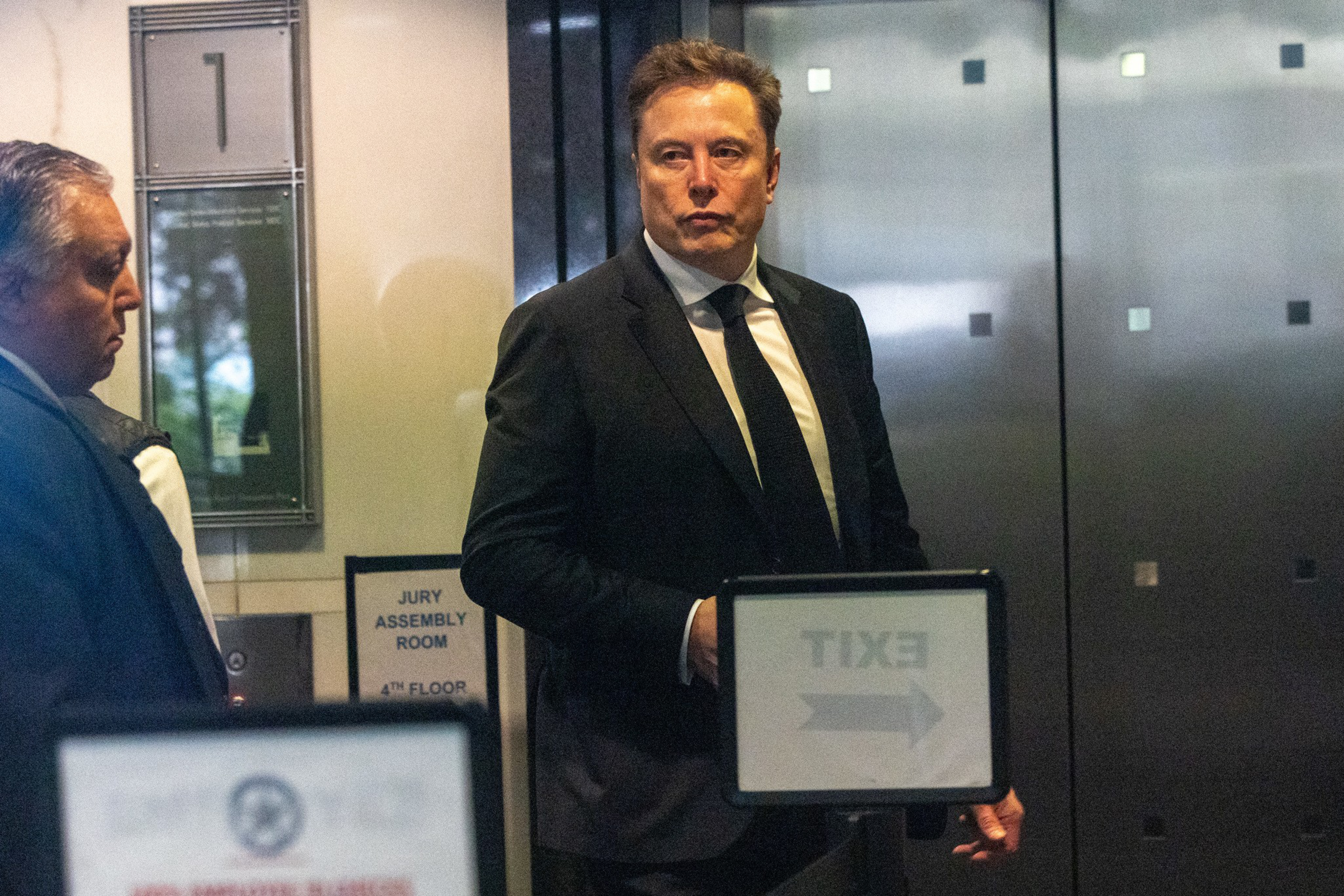 A man in a black suit and tie stands near an elevator with signs indicating a jury assembly room and an exit direction. Another man is nearby.