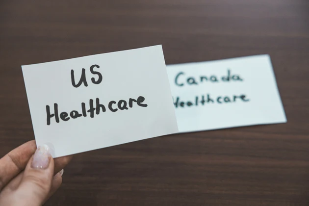 Hand holding two white cards with handwritten text comparing healthcare systems. The foreground card reads US Healthcare while the background card shows Canada Healthcare