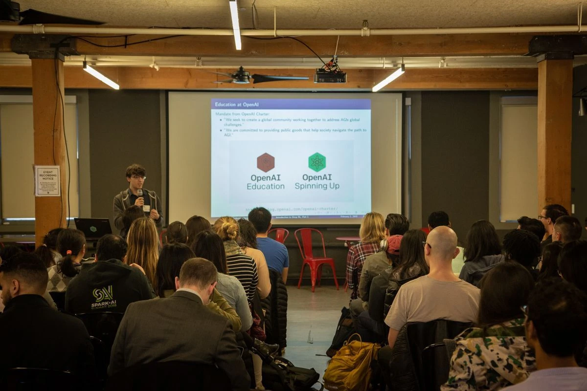 Person speaking into a microphone in front of a room with a live audience