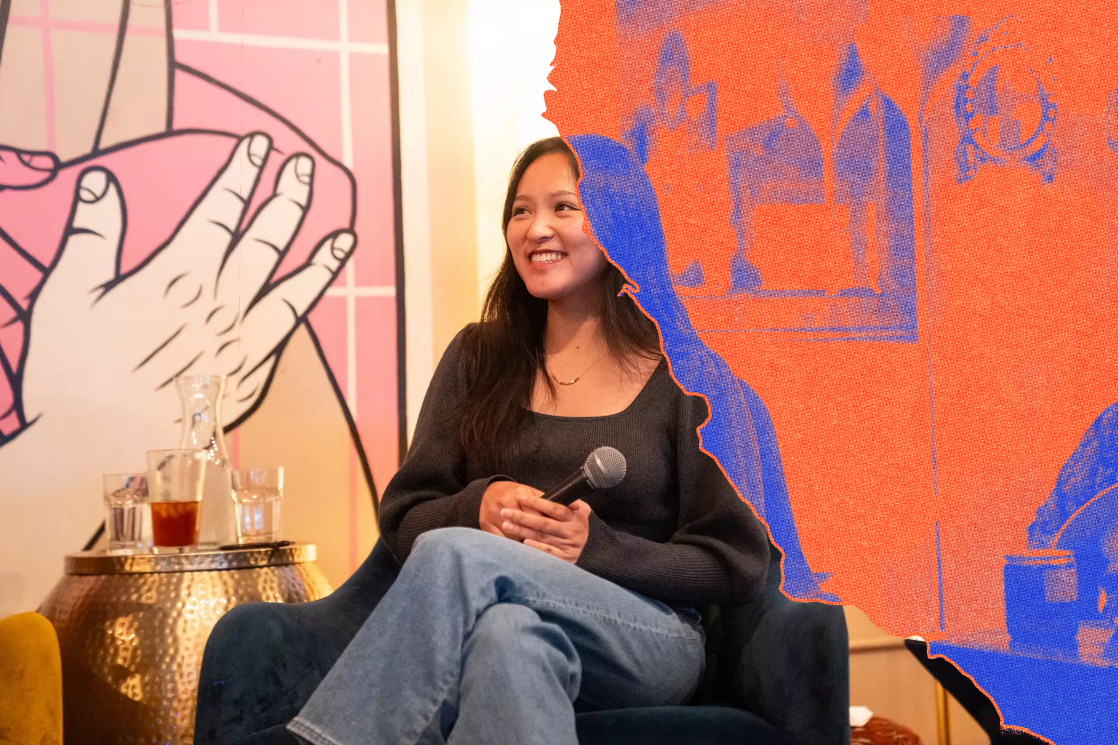 A smiling woman holds a microphone, sitting cross-legged in a chair, with a large colorful hand illustration and drinks on a metallic table nearby.