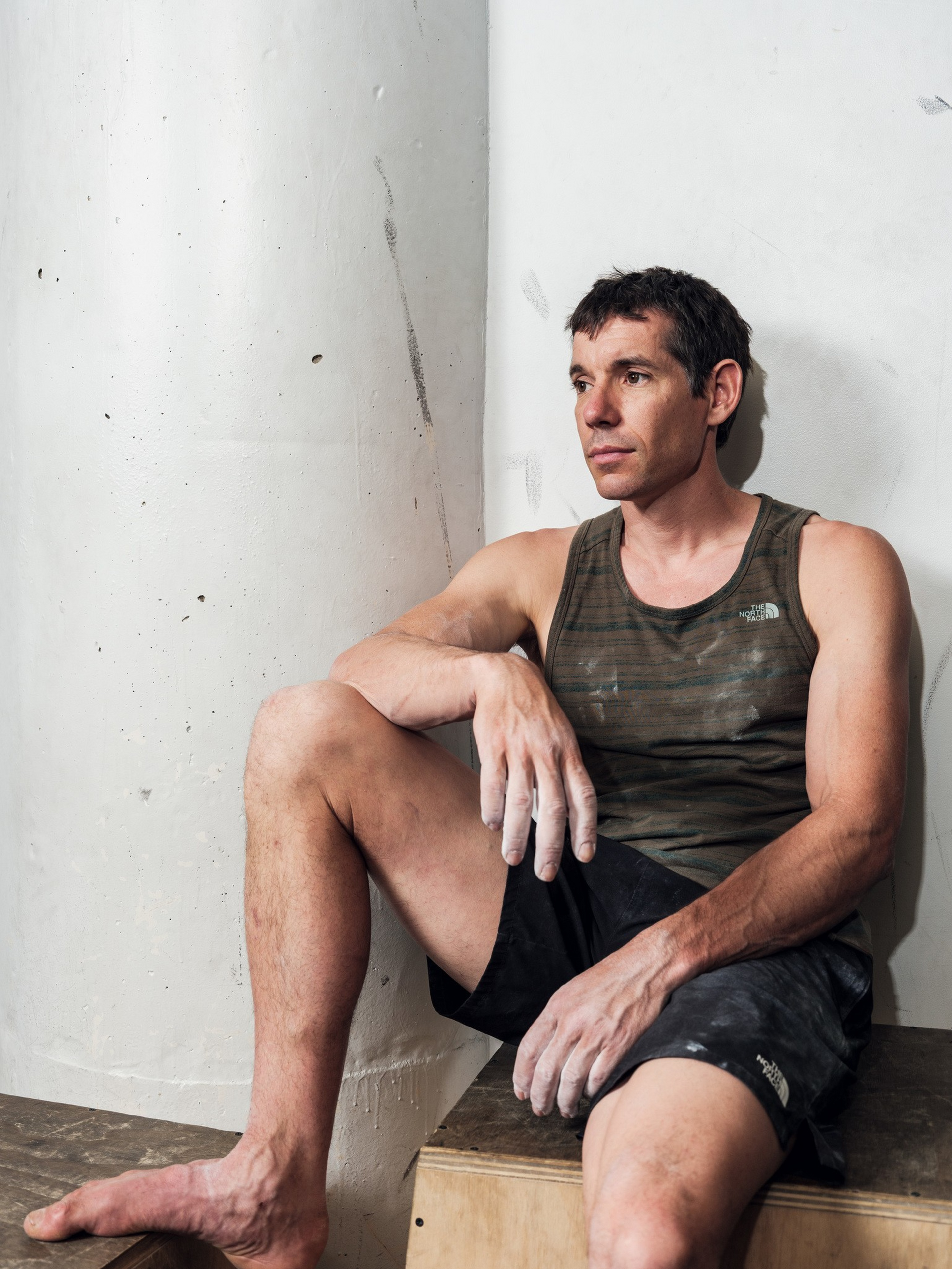 A man in a sleeveless shirt and shorts with chalky hands sits barefoot on a wooden box, leaning against a white wall, looking thoughtfully to the side.