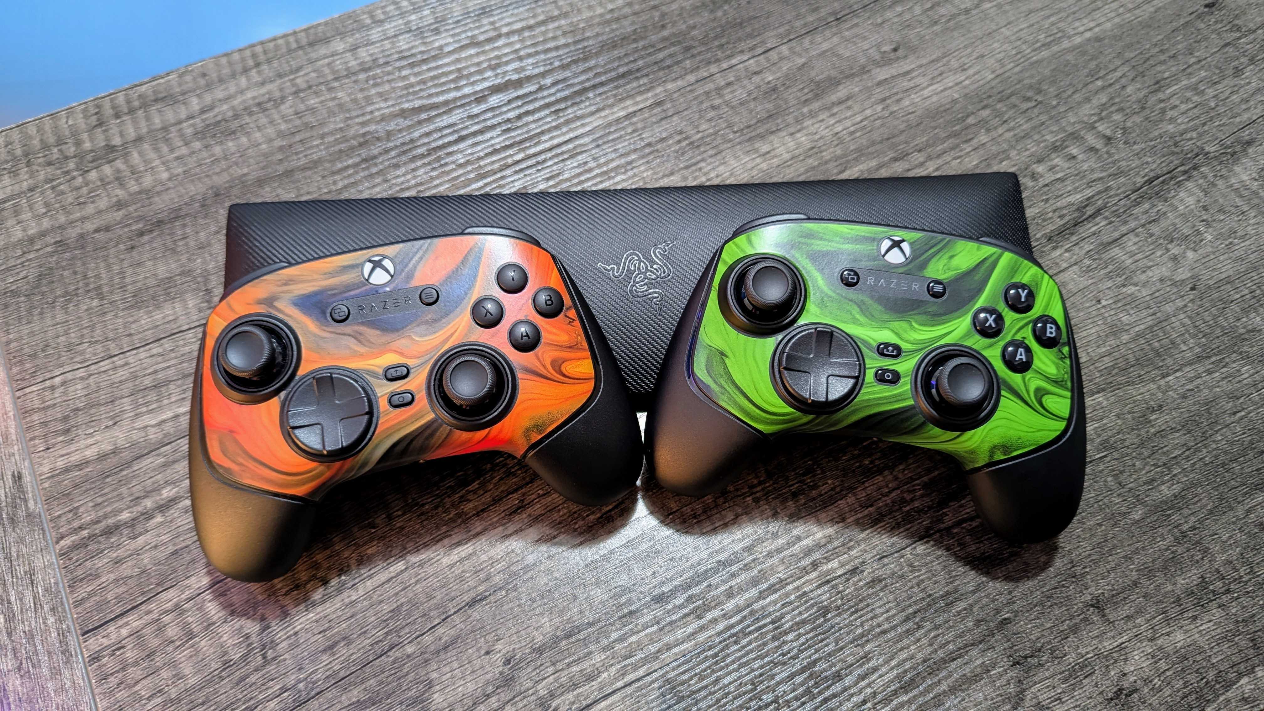 Two Xbox controllers with vibrant swirled designs rest on a wooden surface. One is orange and black, the other green and black, both with a sleek, modern look.