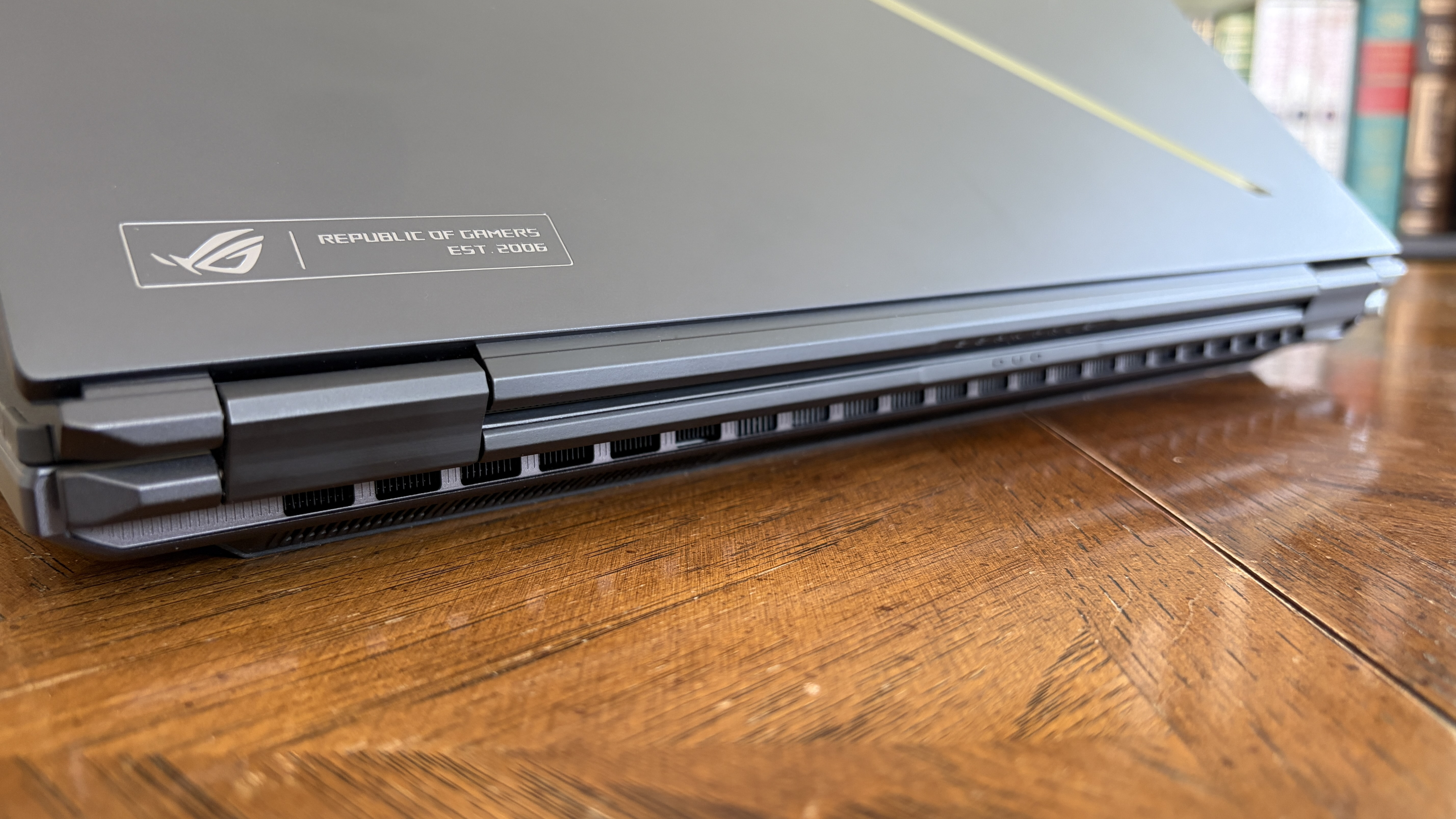 The image shows a closed gaming laptop with a "Republic of Gamers" logo on the lid. It's placed on a wooden surface, with books blurred in the background.