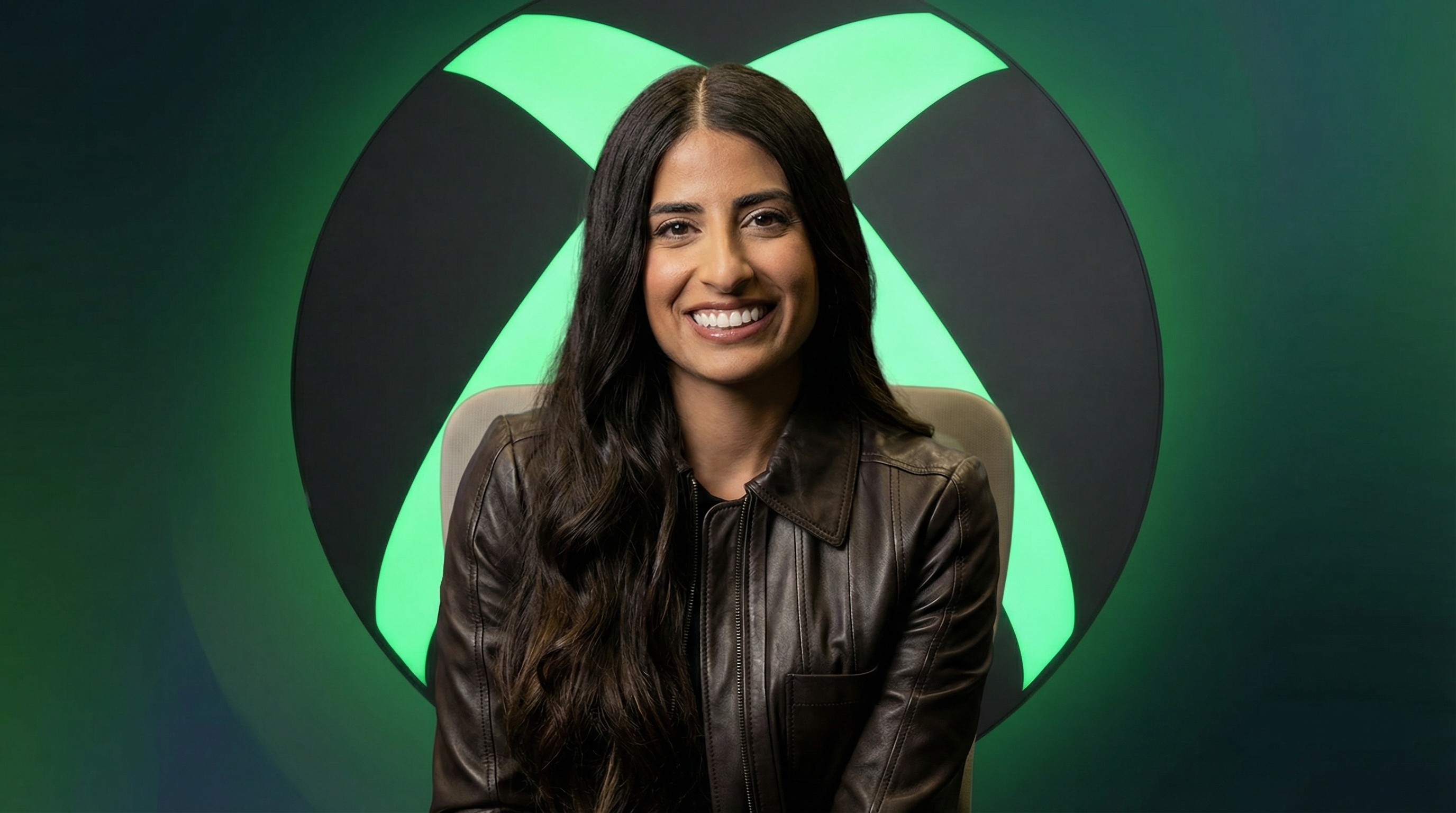 A composited image of Asha Sharma seated in front of a large illuminated Xbox logo on a clean gradient background.