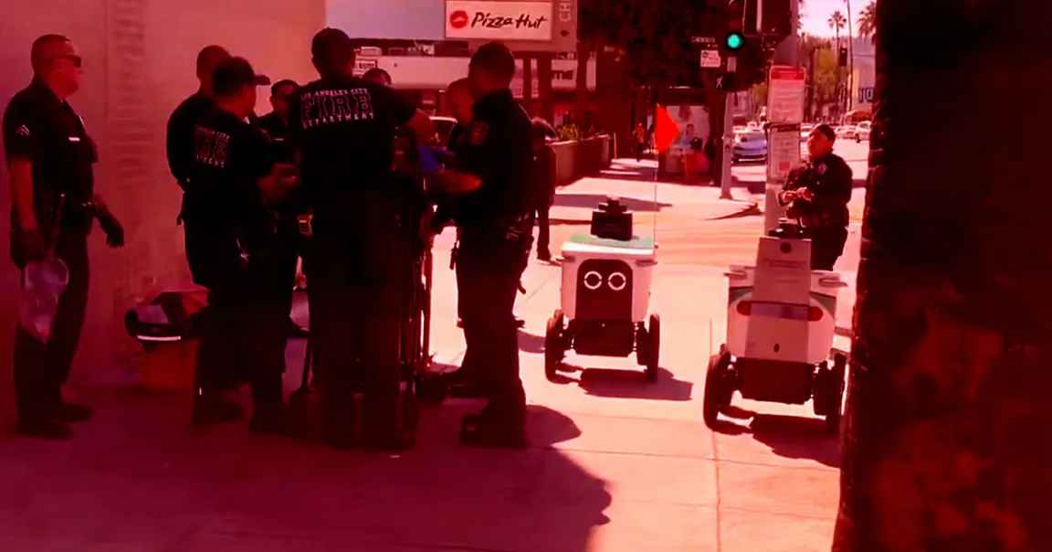 A group of firefighters and police officers are gathered on a sidewalk near a Pizza Hut sign. Two small delivery robots with wheels and flags are on the sidewalk nearby. The scene appears to be in an urban area with traffic lights and cars in the background.