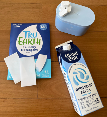 Envelope of laundry detergent sheets, blue tin with dishwasher pods sitting on top, and carton of dish soap on hardwood floor