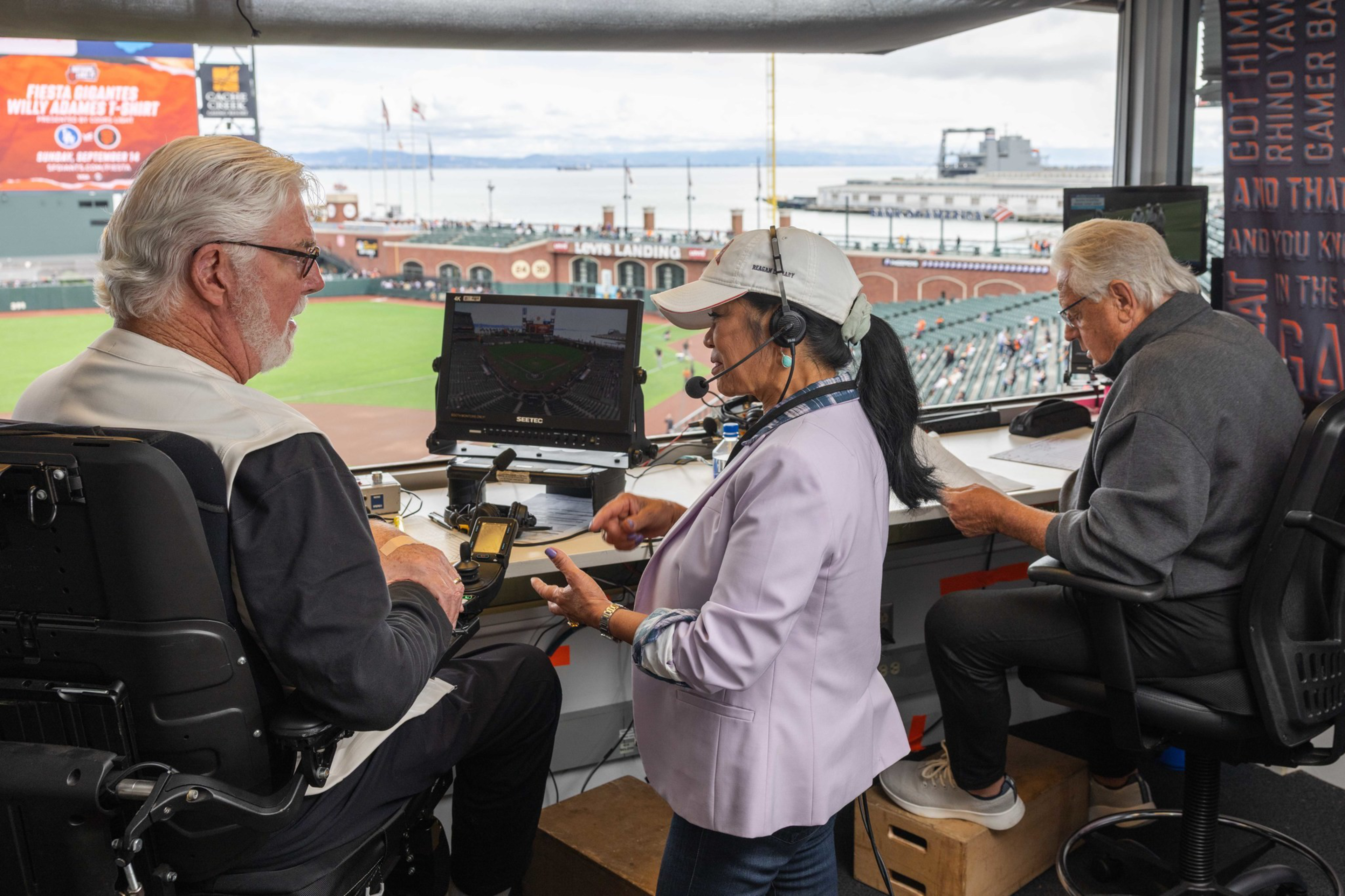Three sports commentators are in a booth overlooking a baseball field; one woman with a headset is talking to two older men seated with equipment.