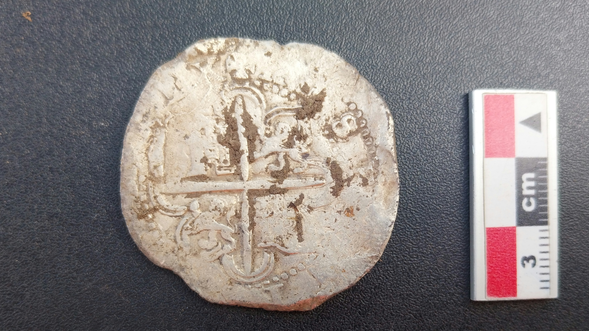 A close up of a silver coin, with small carvings in it. It sits on a black surface with a red and white vertical ruler to its right.