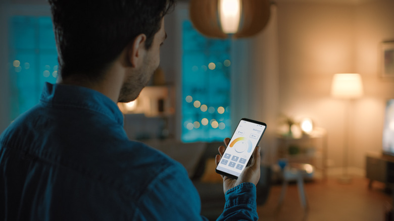 A man holding a smart phone displaying an app to control smart lights with a blurry living room background.