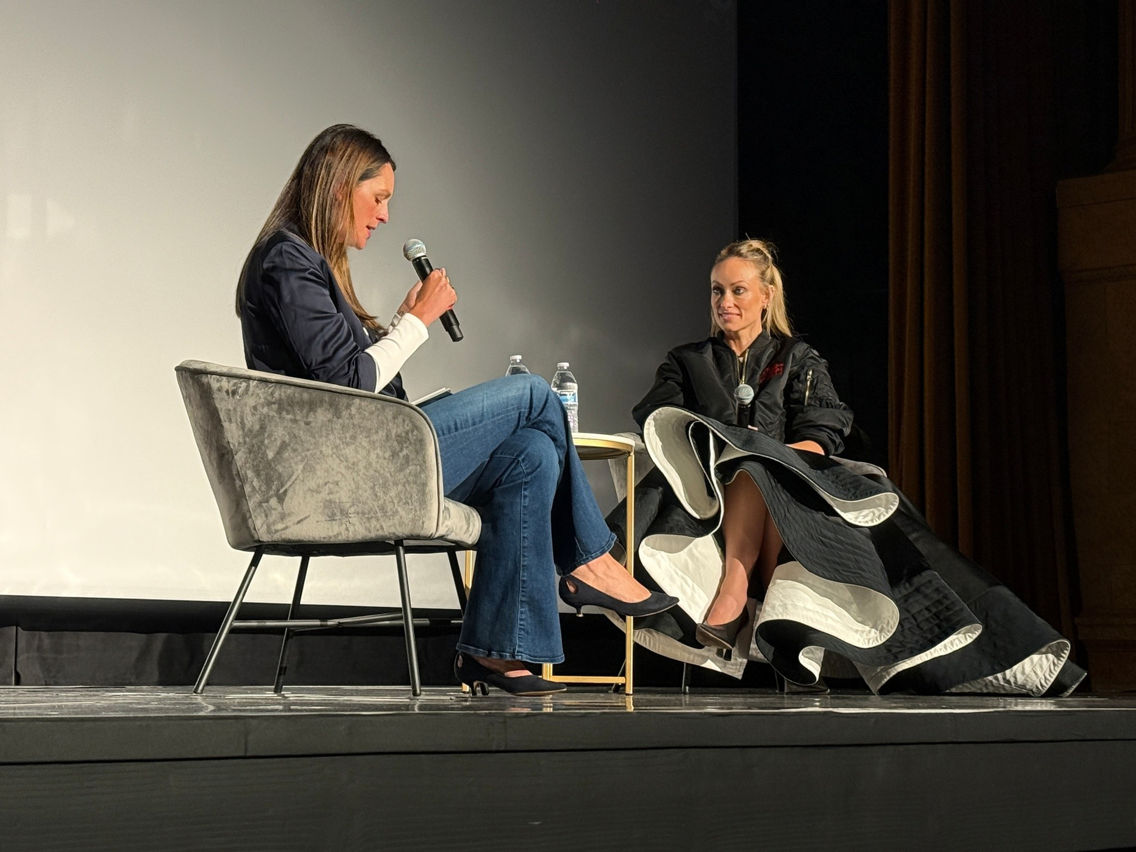 Two women sit on stage in armchairs; one holds a microphone and reads from a paper, the other listens, wearing a dramatic black and white dress.