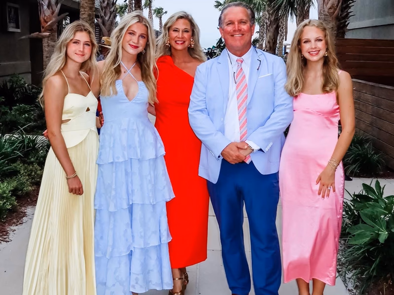 A family of five stands together between palm trees.