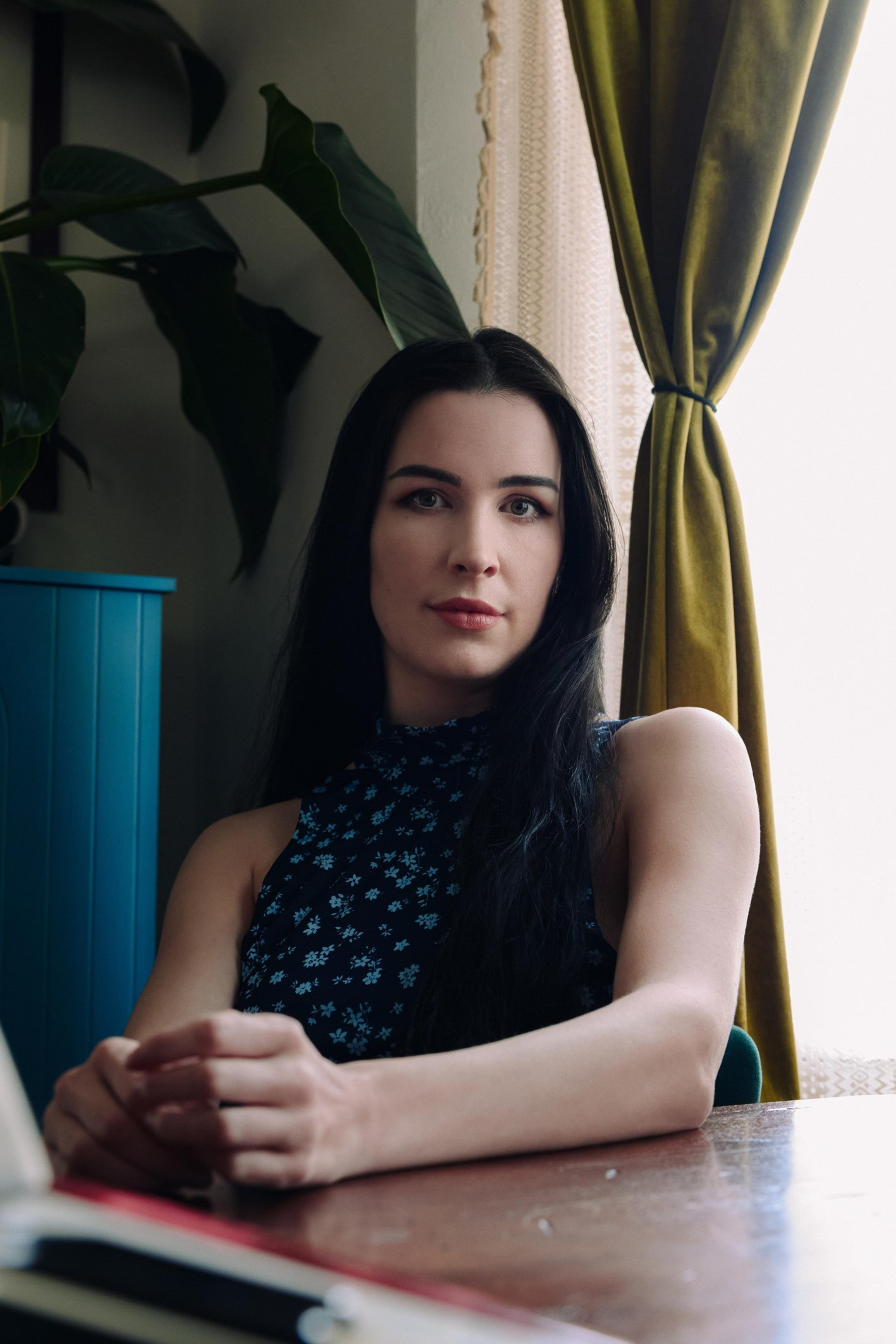 A woman with long dark hair sits at a wooden table, wearing a sleeveless dark top with a floral pattern, with a green curtain and plant in the background.