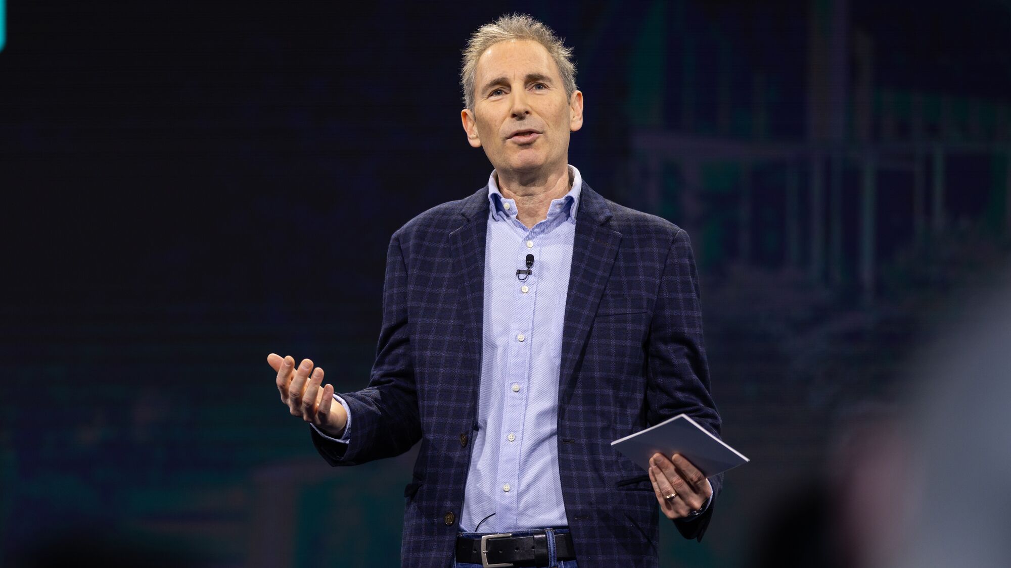 Amazon CEO Andy Jassy wearing a plaid blazer presenting on large stage in front of Amazon employees.