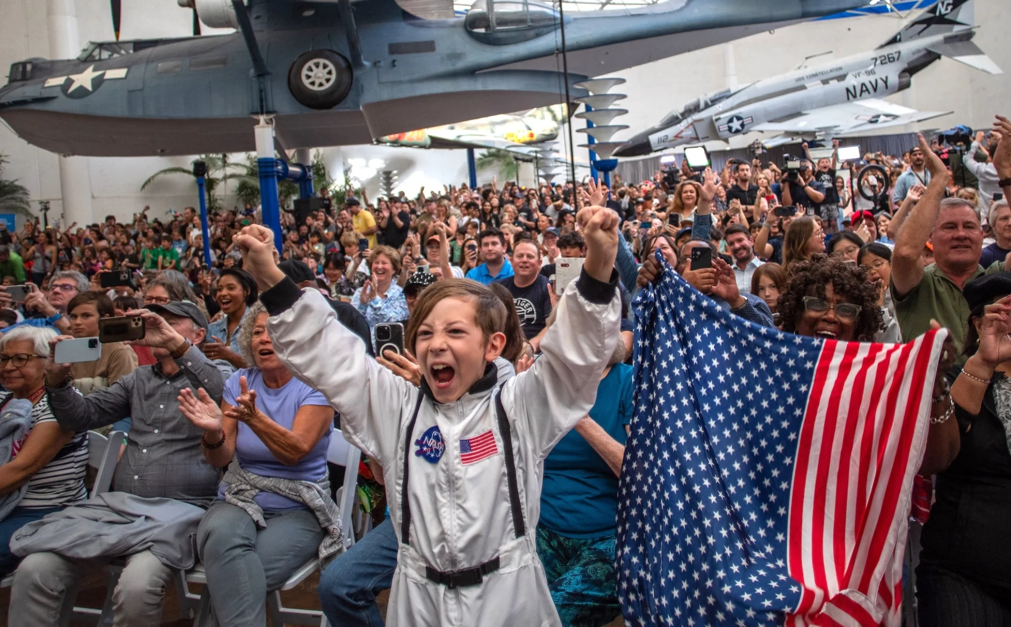 A young boy wears an astronaut costume next to a woman waving a flag as they watch a live broadcast of the return of the Artemis II