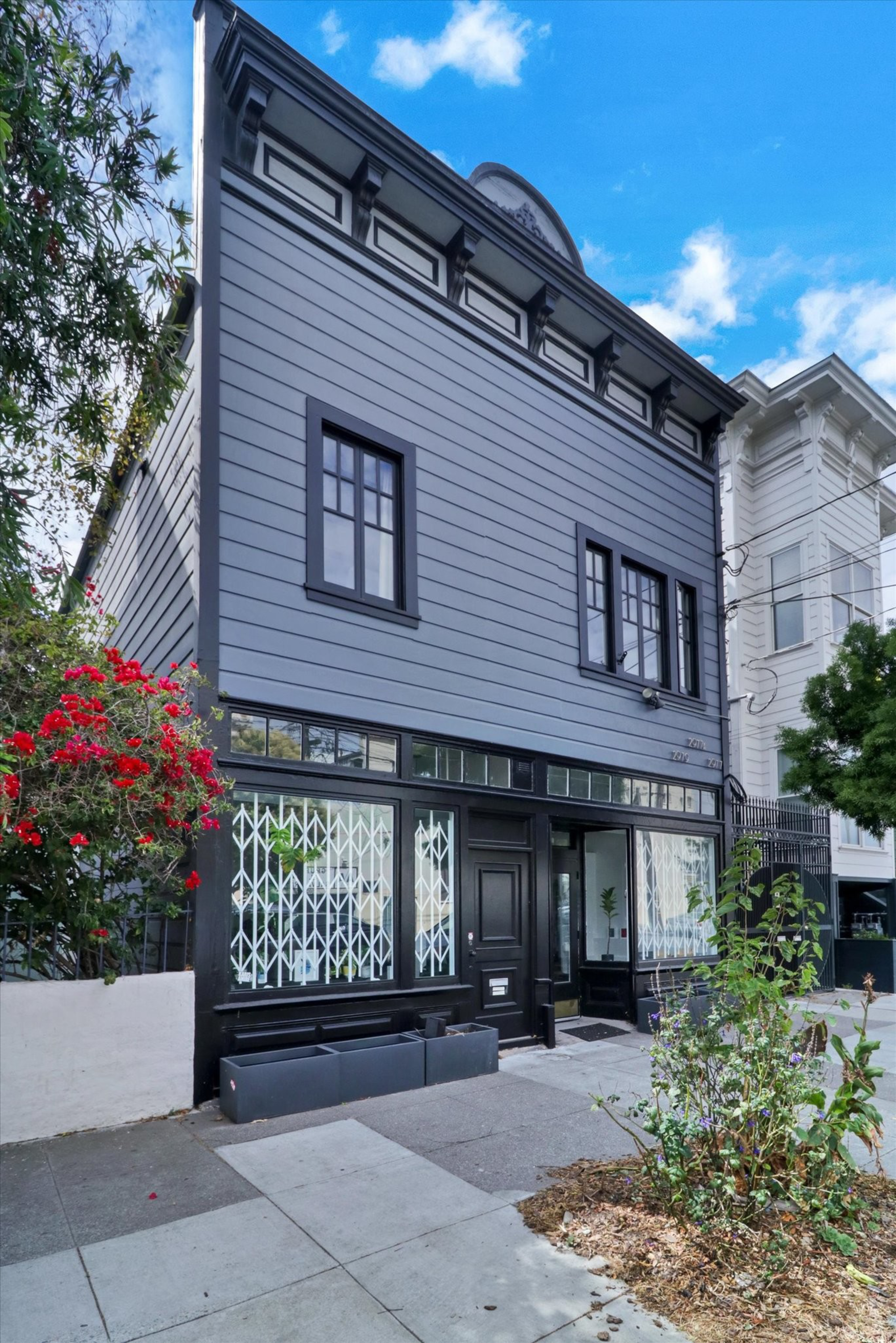 A two-story gray building with black trim has large windows on the ground floor with white security bars, red flowers on the left, and a small plant in front.
