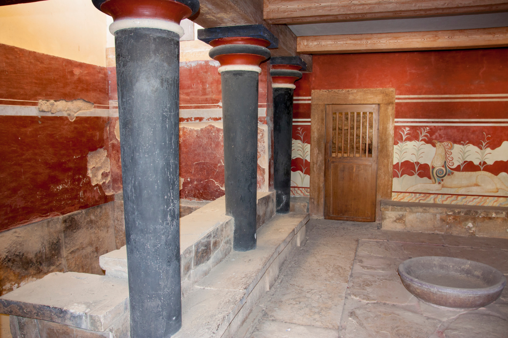 Inside an ancient building, scarlet red walls are seen with a wooden door built into the far wall. To the left are black pillars.
