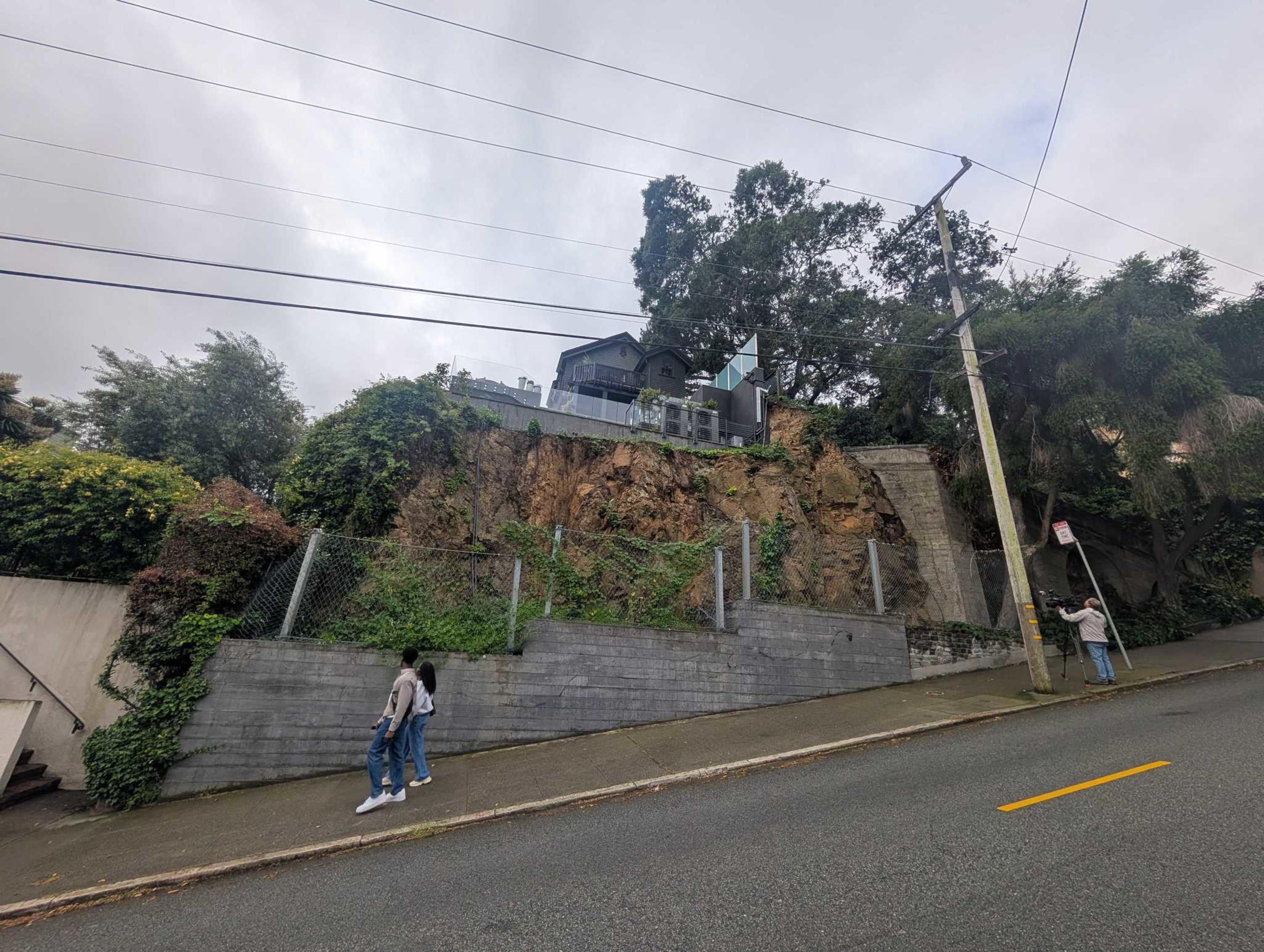 A steep sidewalk runs along a road with a yellow line, bordered by a rocky hill with greenery and a house atop, while three people walk or stand nearby.