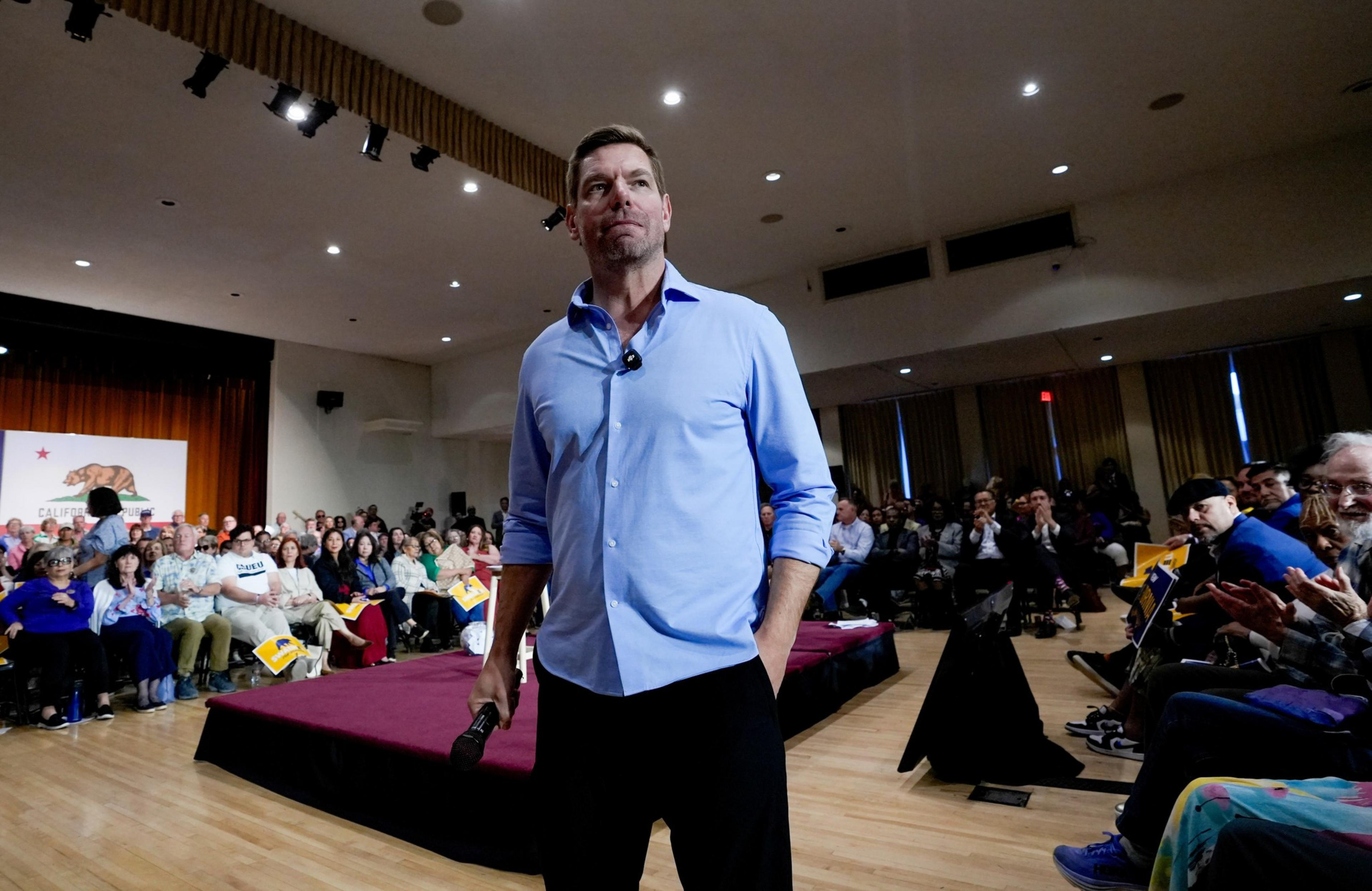 A man in a light blue shirt stands confidently with one hand in his pocket and a microphone in the other, facing an applauding audience in a large hall.