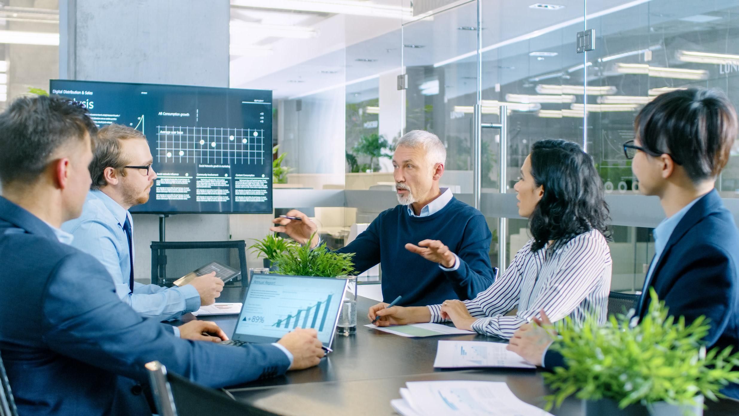 Senior Executive erklärt Vision und Potenzial des Unternehmens für seine Mitarbeiter. Sie sitzen am großen Tisch im Sitzungszimmer. Der Fernsehbildschirm an der Wand zeigt das Unternehmenswachstum.