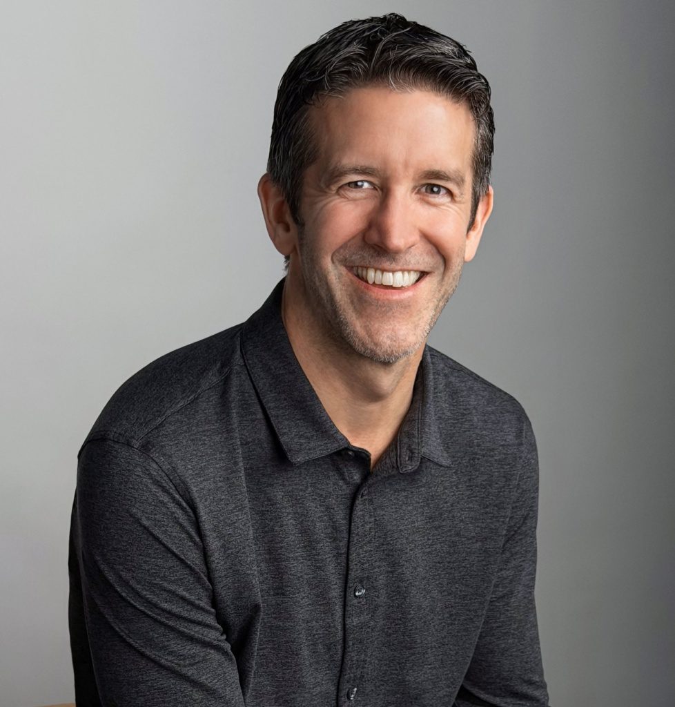 Portrait of Apple executive John Ternus smiling against a plain grey background.