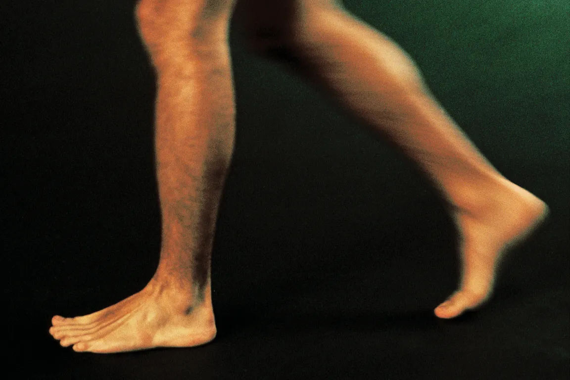 Two bare legs and feet of a person walking or running, with one foot flat on the ground and the other foot raised on the toes, set against a dark background.