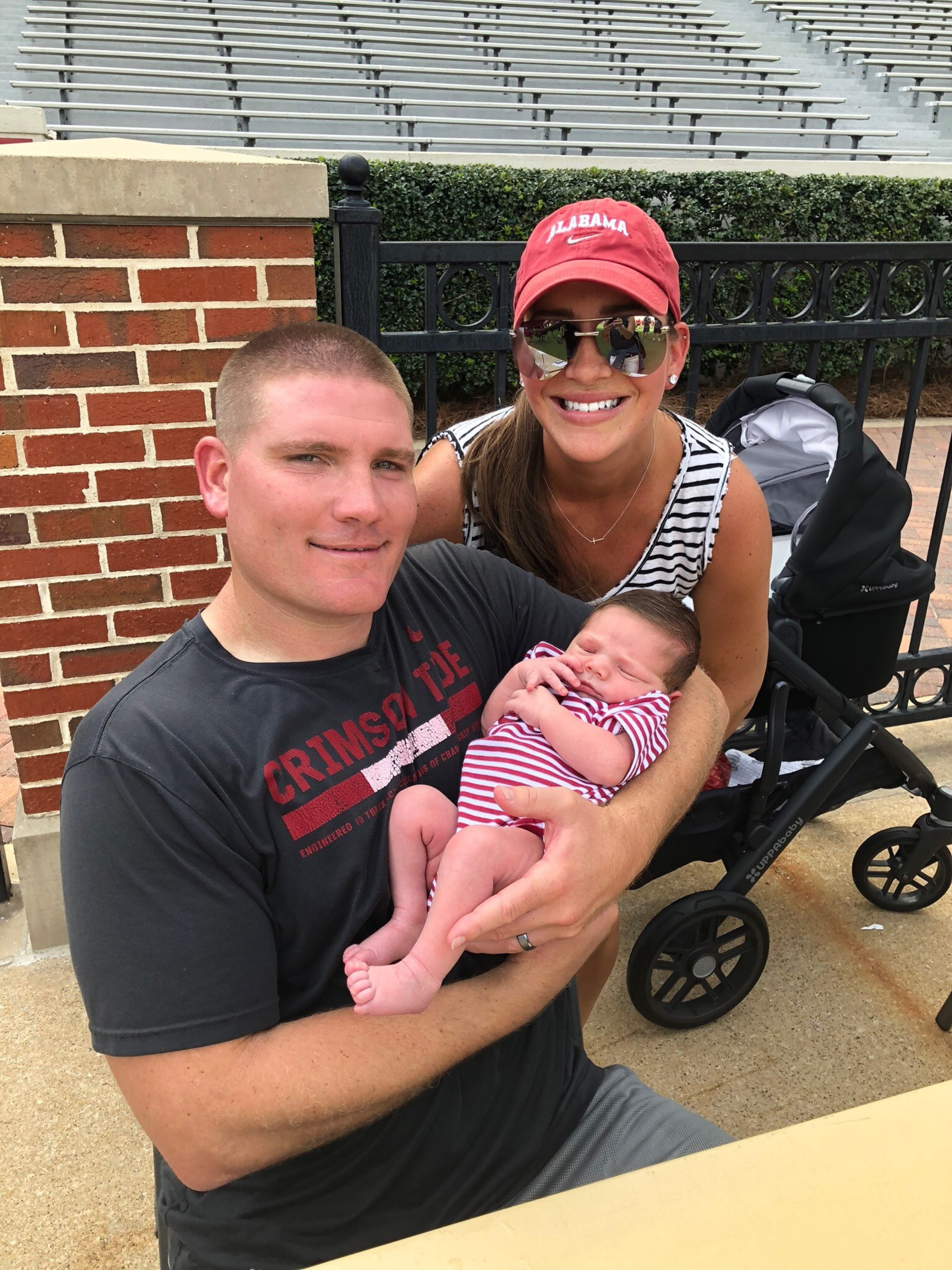 A man wearing a Crimson Tide shirt holds a sleeping baby in a striped outfit, while a woman in an Alabama hat and sunglasses smiles beside them.