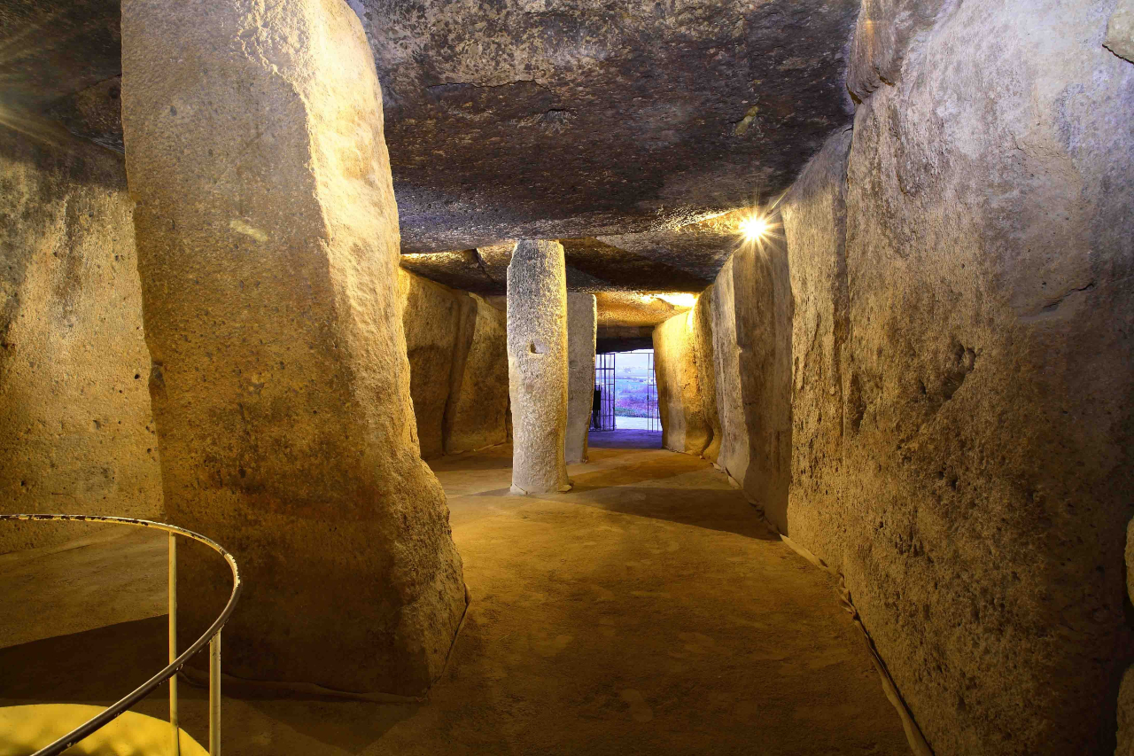 A view into a rectangular shaped stone tunnel with large pillars to the left holding up the ceiling.