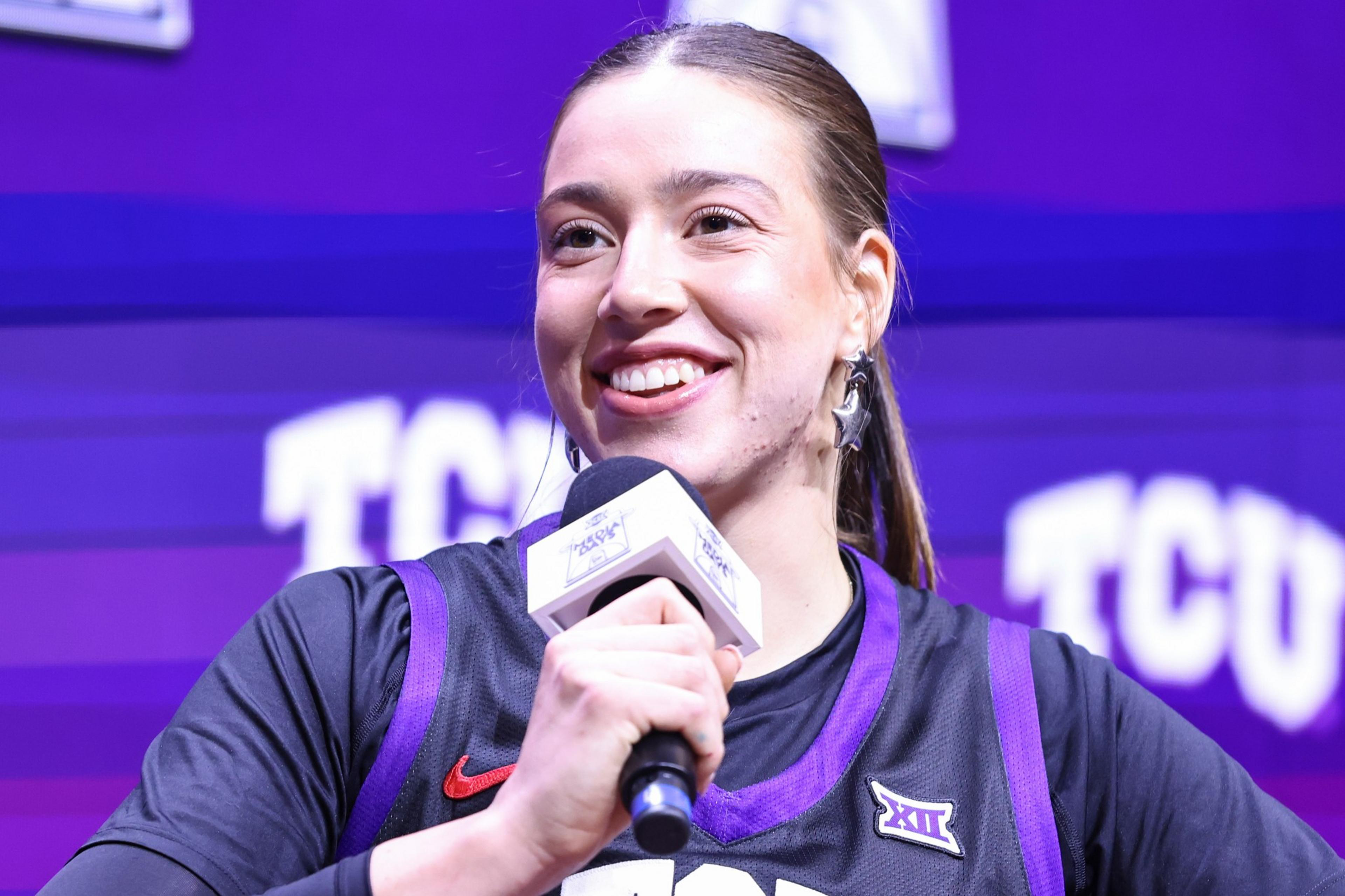 A young woman in a black and purple sports jersey smiles while holding a microphone, with a purple TCU backdrop behind her.