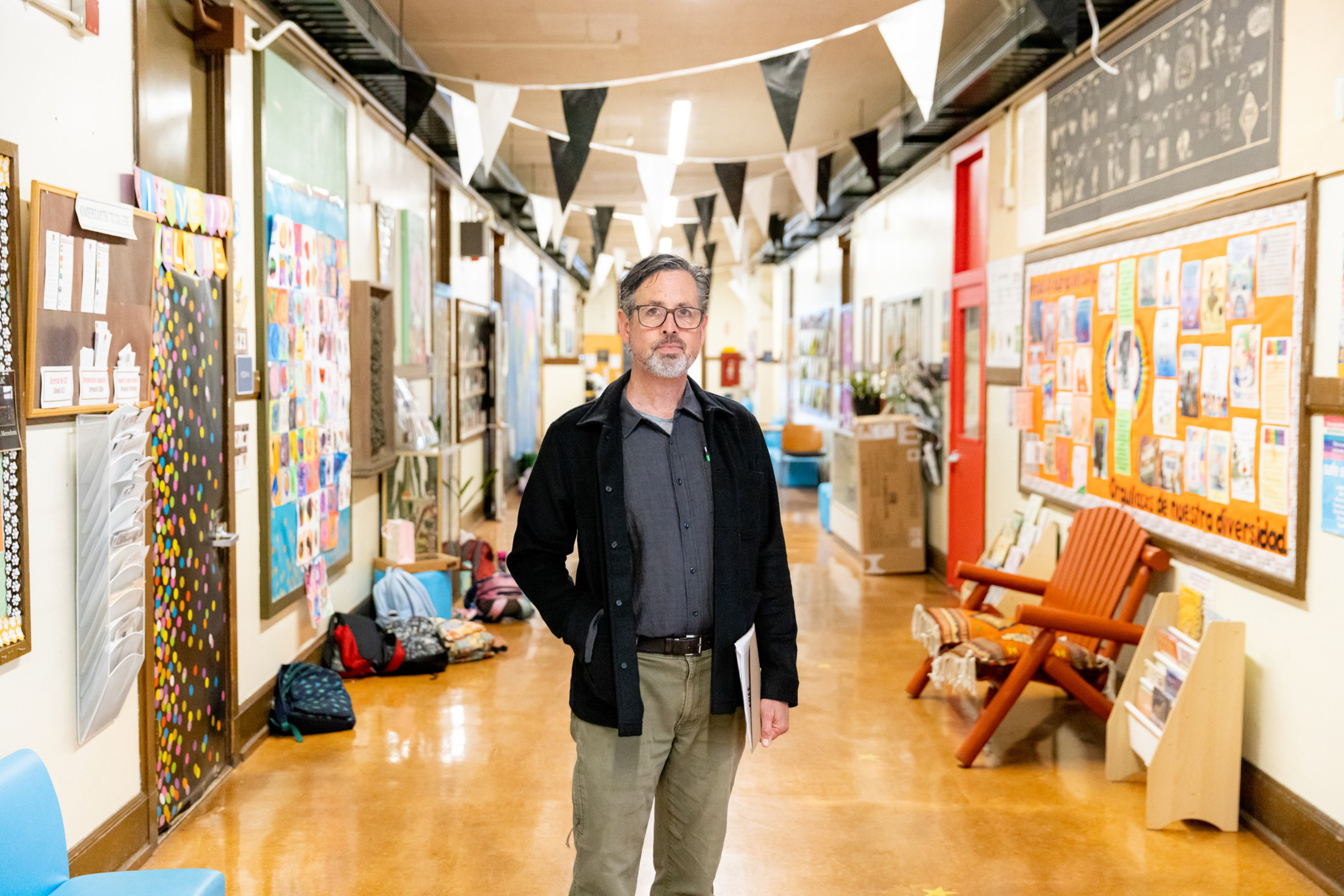 A man stands in a brightly lit school hallway with colorful bulletin boards, backpacks on the floor, and a wooden chair to one side.