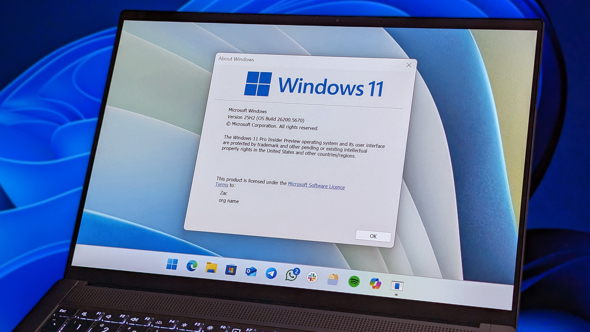 A laptop screen displays the "About Windows" dialog box for Windows 11, showing version details. The desktop has several icons, set against a blue-toned background.