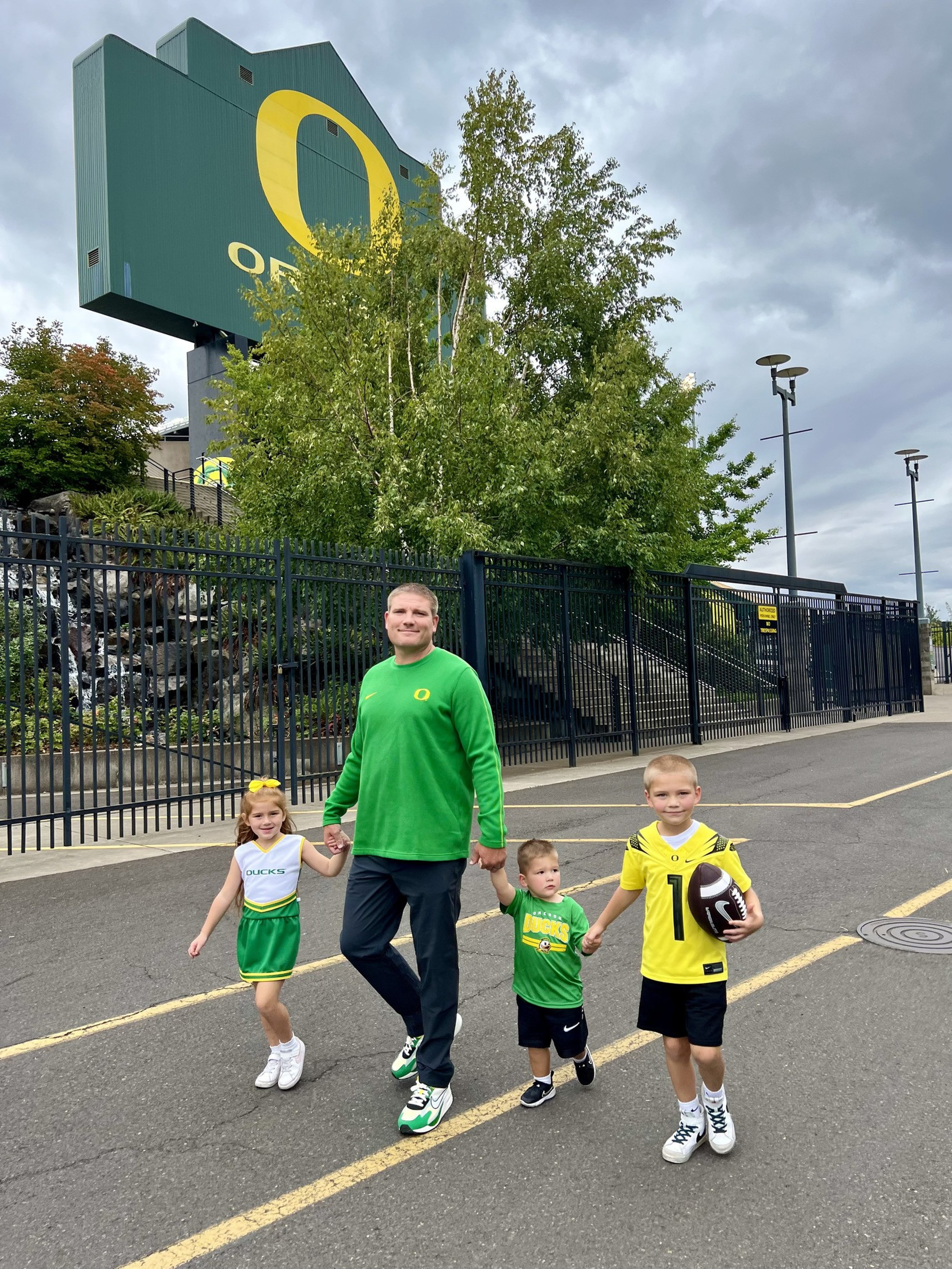 A man and three children, all dressed in green and yellow Oregon Ducks gear, stand in front of a large “O” sign near a stadium fence under a cloudy sky.