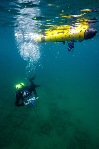 A diver operates below a yellow autonomous underwater vehicle during an underwater human-machine teaming test