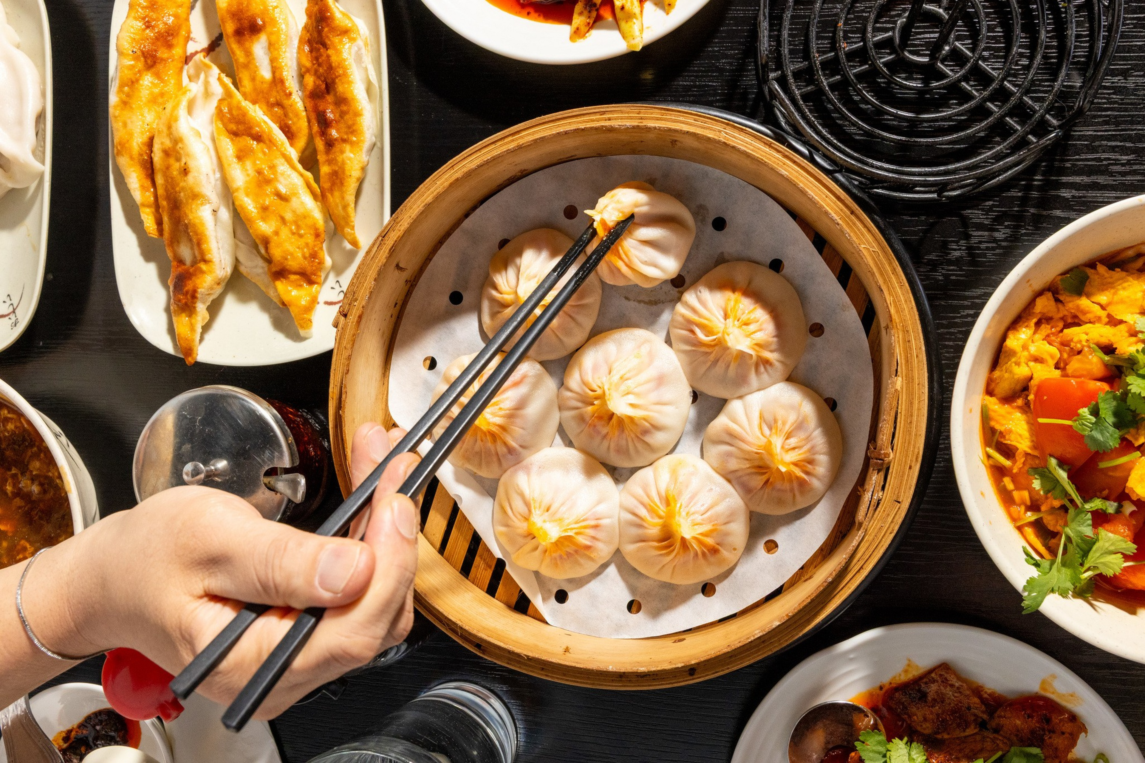 A hand using chopsticks picks up a dumpling from a bamboo steamer surrounded by plates of gyoza and vegetable dishes.