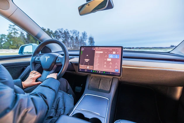 Close-up view of driver holding steering wheel inside Tesla Model 3 electric car with touchscreen displaying vehicle controls.