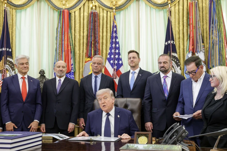 Donald Trump seated at his desk in the Oval Office surrounded by cabinet members.