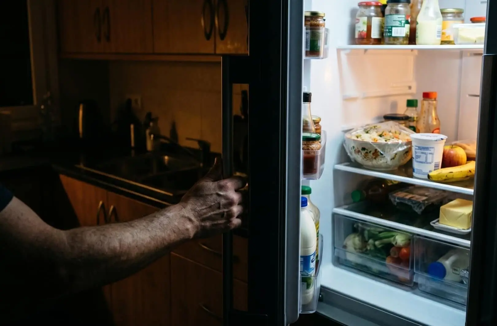 Recreación artística que muestra a una persona abriendo un frigorífico en una cocina doméstica de noche, revelando alimentos bajo la luz interior en contraste con la oscuridad de la habitación. Foto: Nano Banana / Scruzcampillo.